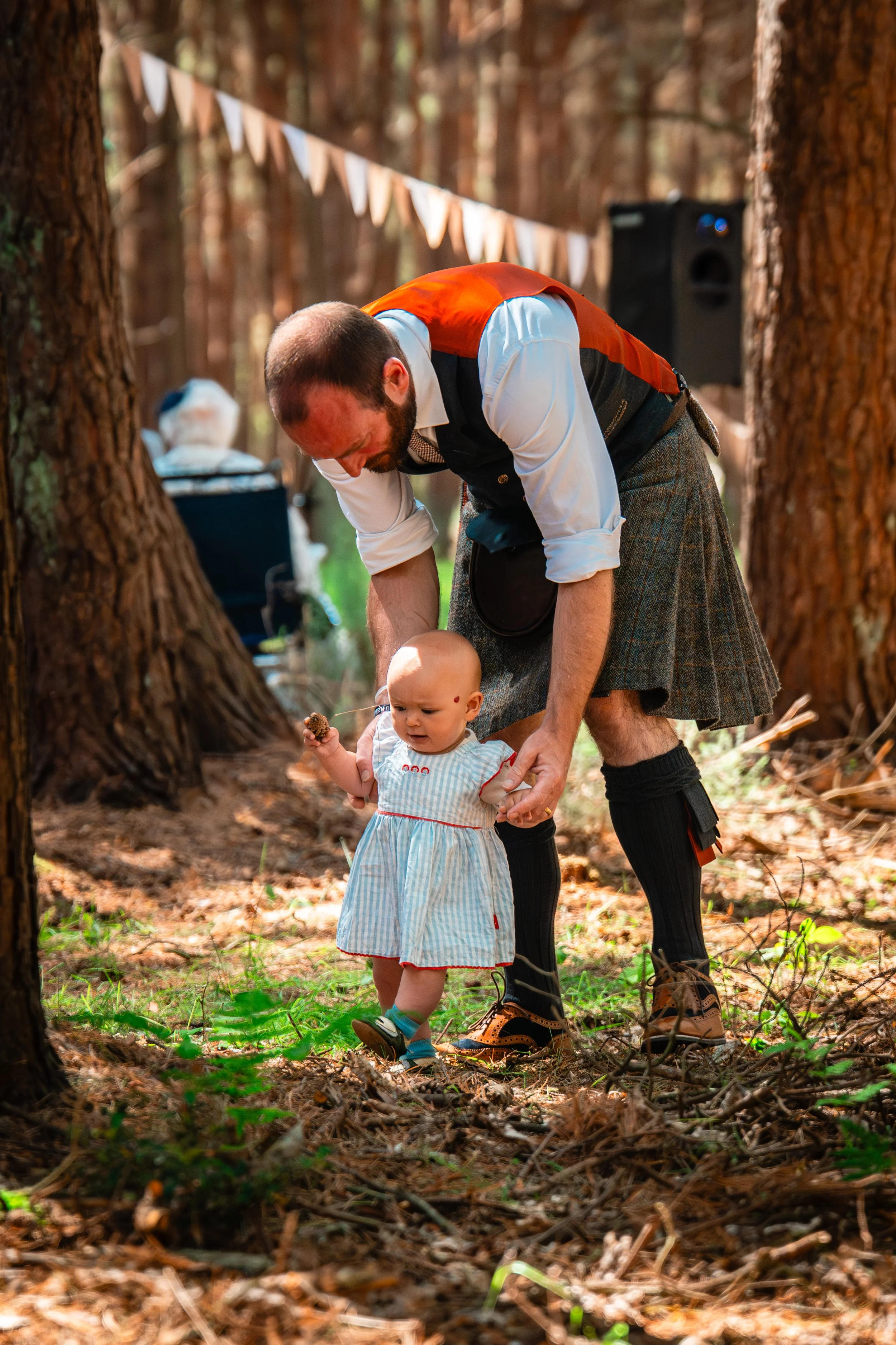 A man dressed in traditional Scottish attire, including a kilt, helping a young child walk in a wooded outdoor setting during a festive event, with a speaker and string flags visible in the background.