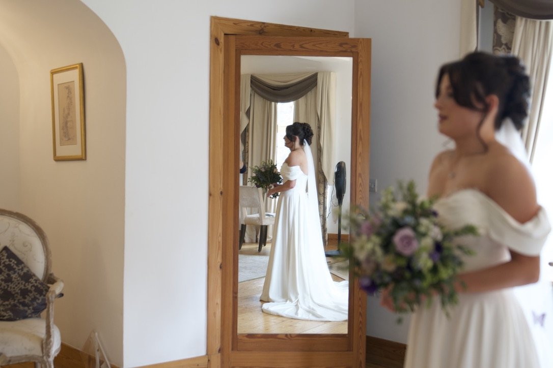 A bride in a white wedding dress holding a bouquet, standing in front of a large mirror that reflects her, a dining room table, and curtains.