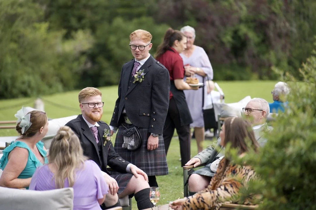 People gathered outdoors at a social event, some sitting and others standing, with trees and greenery in the background.