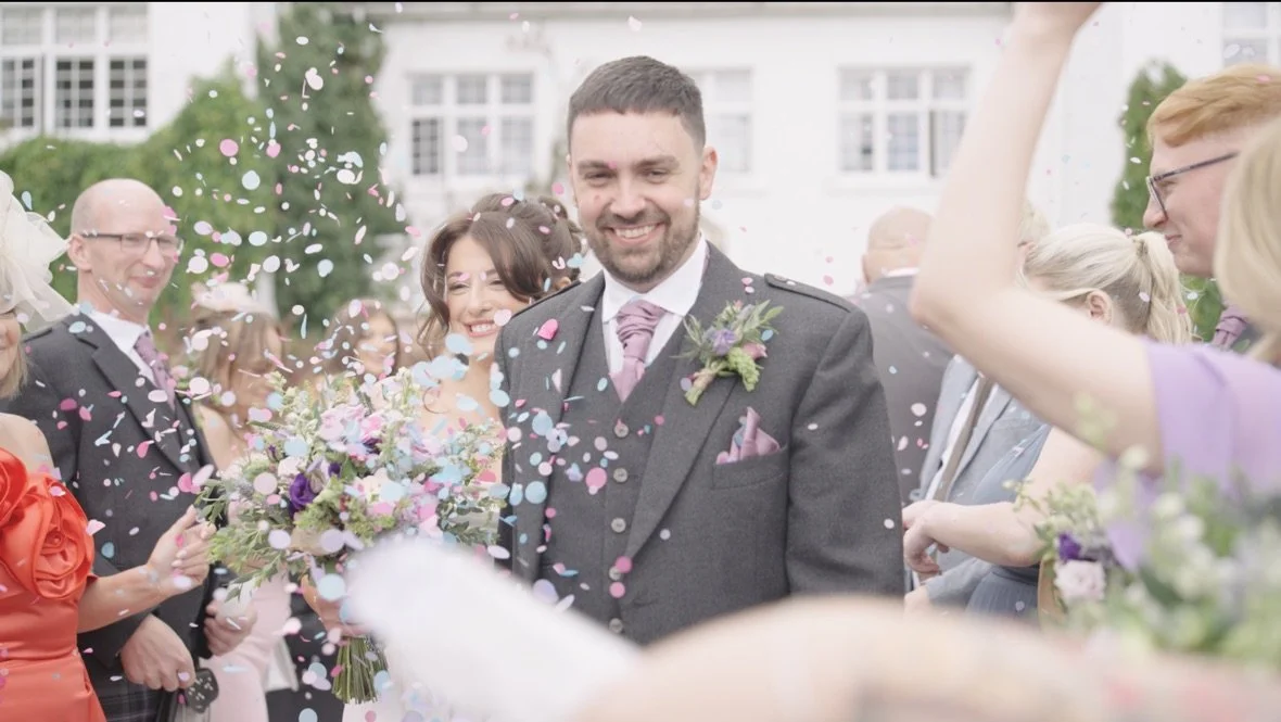 A groom in a gray suit with a pink tie and boutonniere smiling as confetti falls around him at a wedding celebration.