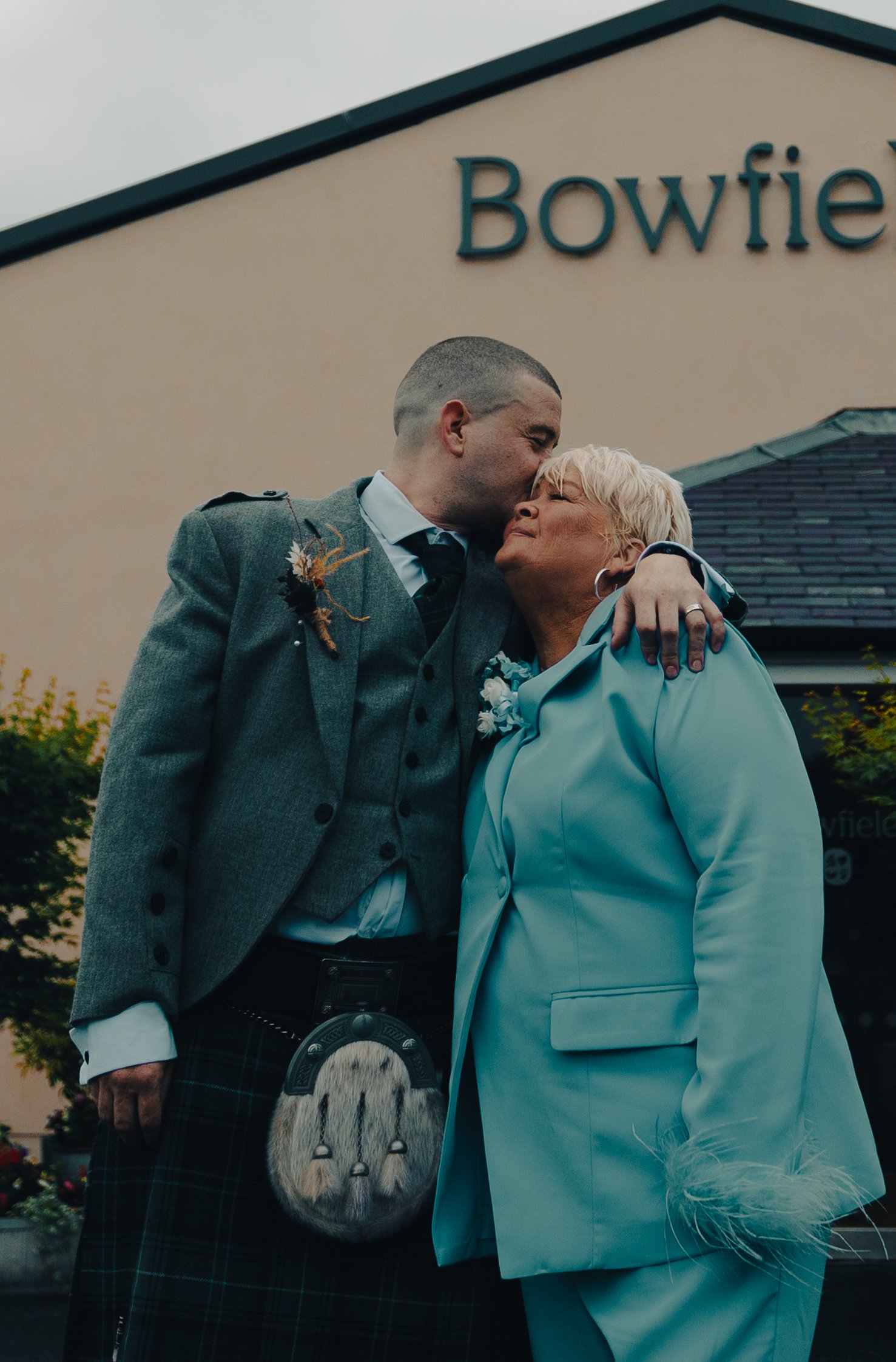 A young man in a grey suit with a tartan kilt and sporran kisses an older woman in a light blue suit outside a building with a sign that says 'Bowfire'.