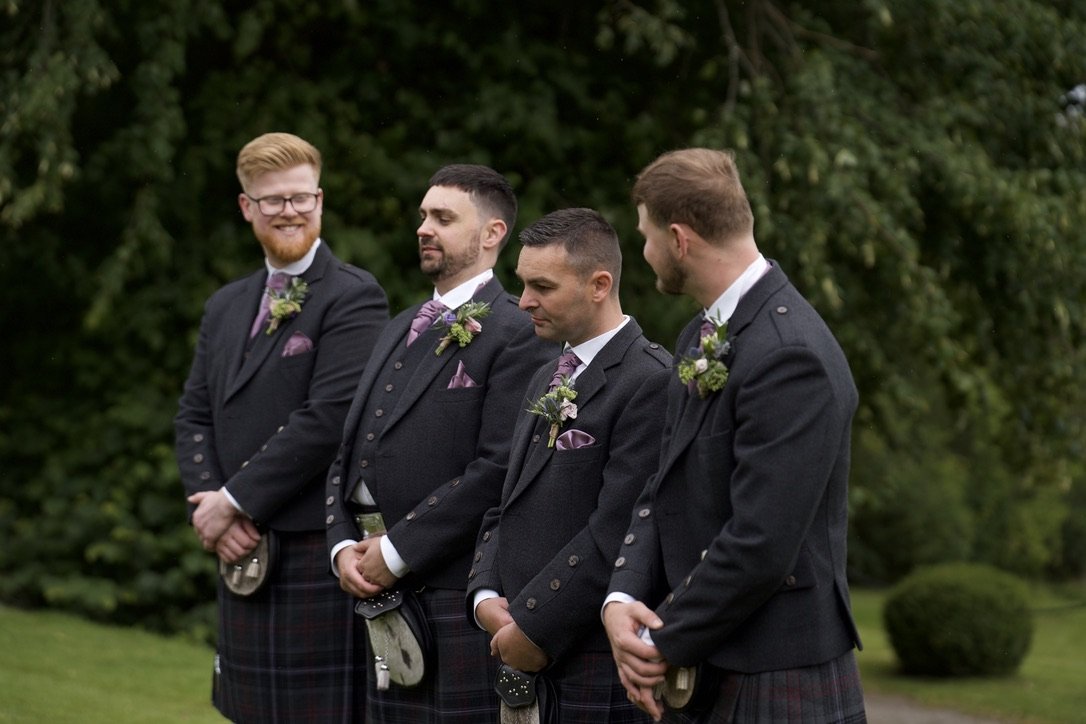 Four men dressed in suits and kilts standing outdoors with greenery in the background, possibly at a wedding or outdoor event.