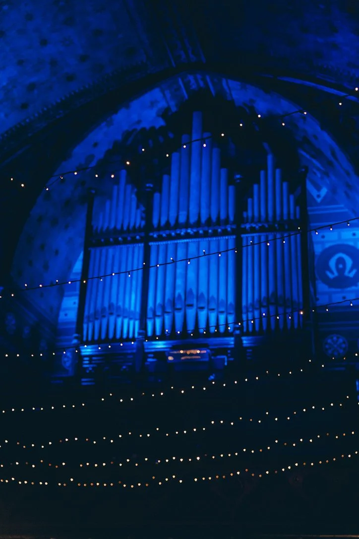 A dark image showing a large organ with illuminated pipes in a church or concert hall, decorated with string lights.