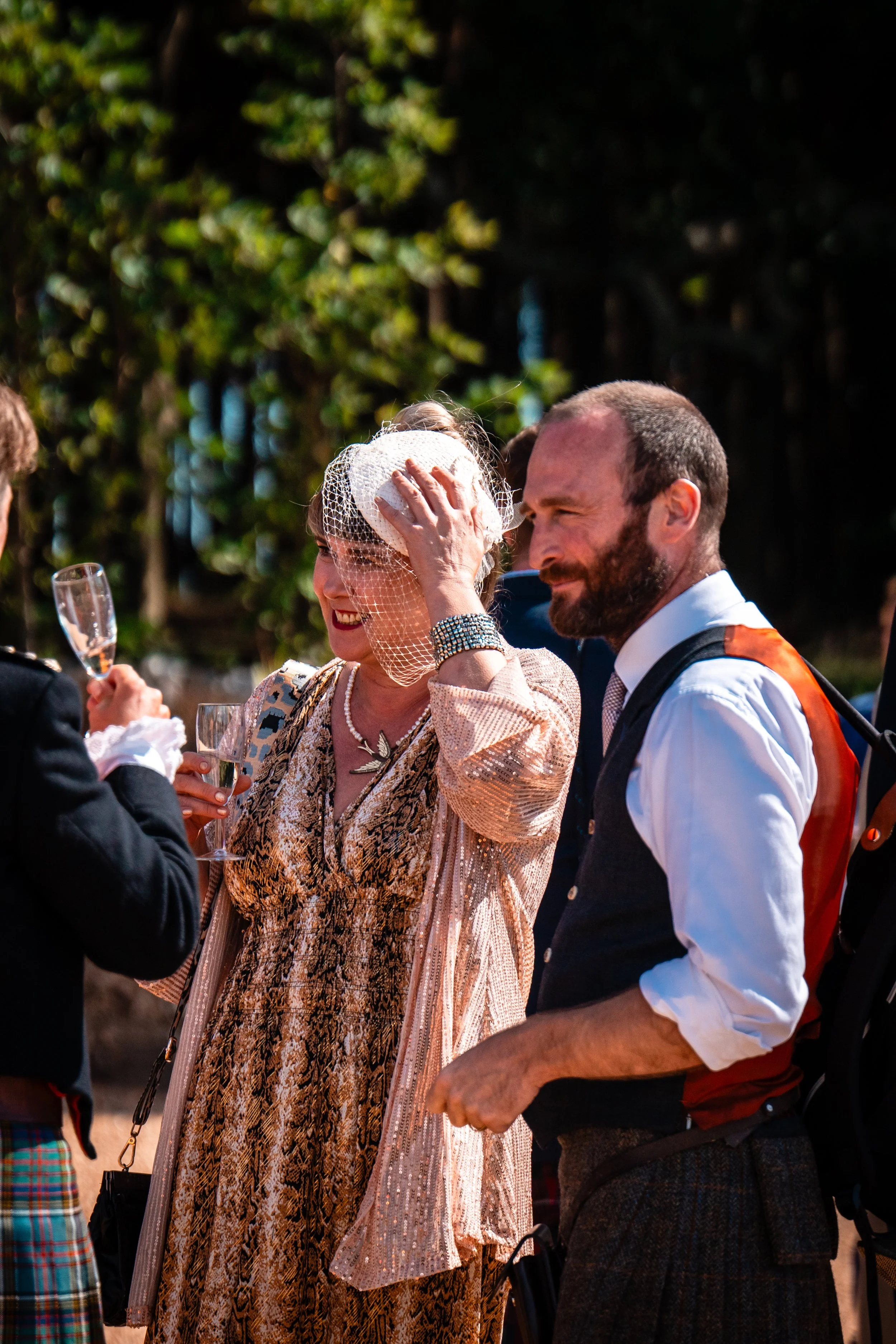 A woman in a vintage dress with a fishnet hat adjusts her hat while holding a glass of wine, surrounded by people outdoors with trees in the background.