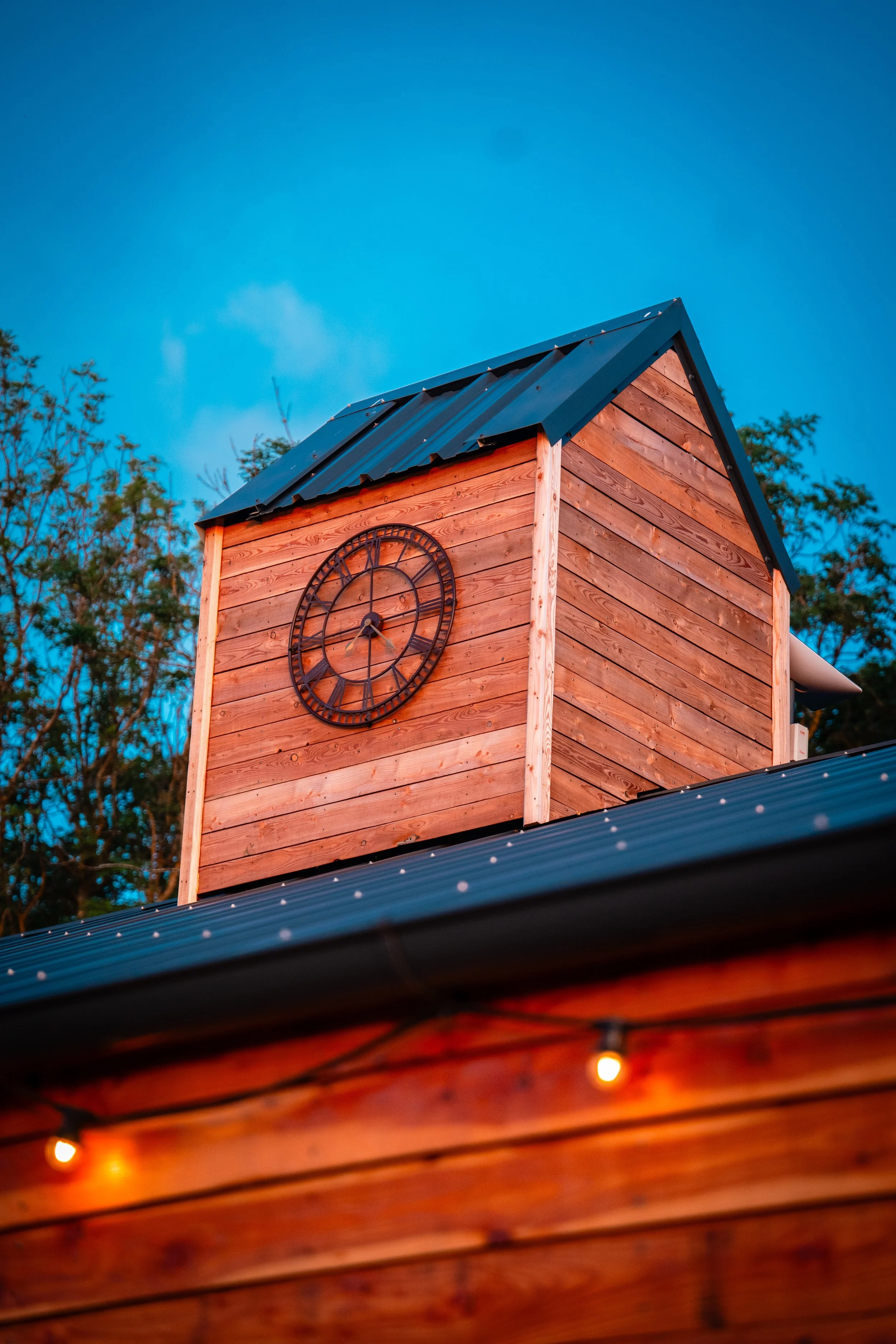 A wooden clock tower with a black metal roof and a round clock face mounted on the front, outdoors during evening twilight.