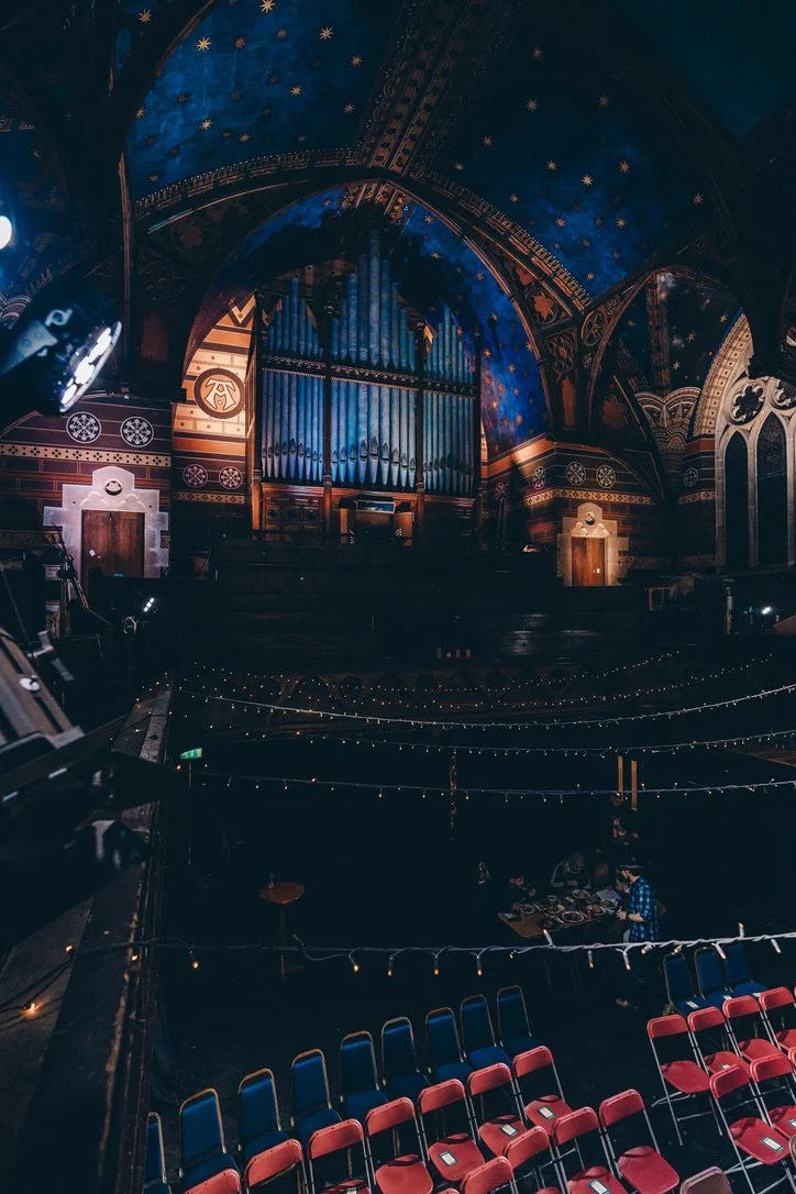 Inside a historic concert hall with an ornate, dark blue ceiling decorated with stars, featuring a large pipe organ on stage, red cushioned chairs arranged in rows, and string lights hanging across the seating area.