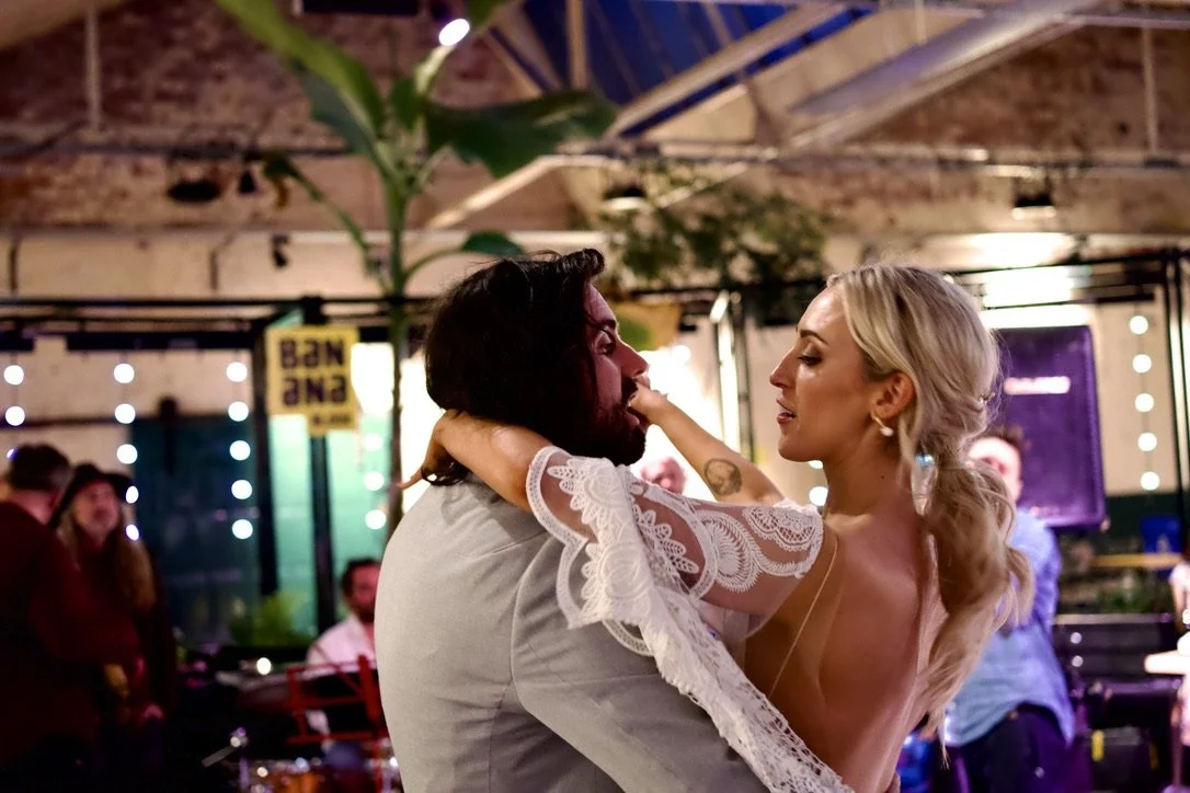 A couple dances closely in a lively indoor setting with colorful lights and people in the background.