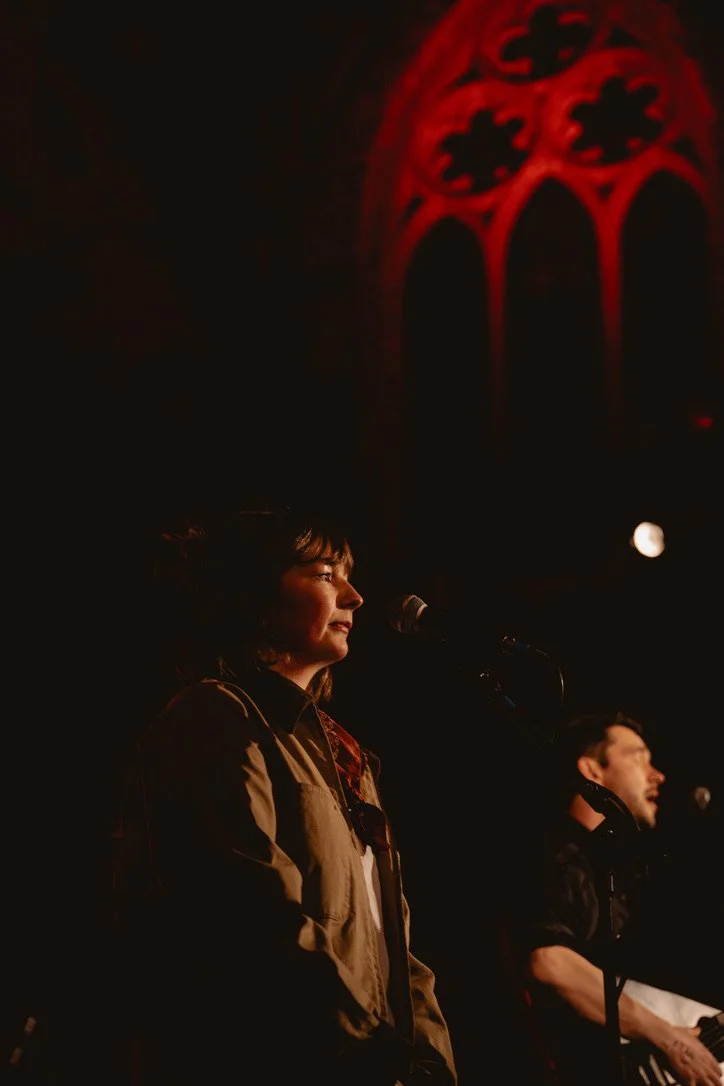 A woman and a man are standing on stage with microphones, with a red and black gothic stained glass window in the background, under low stage lighting.