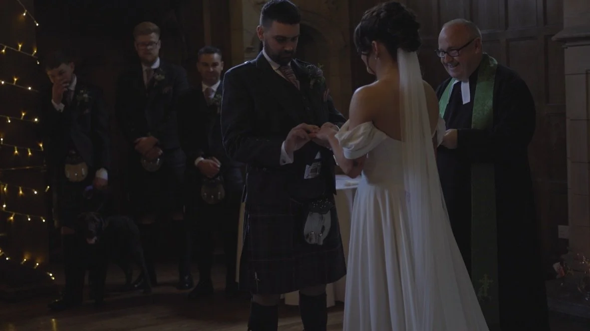 A wedding ceremony with a bride and groom exchanging rings in front of a priest, with groomsmen and a dog in the background.