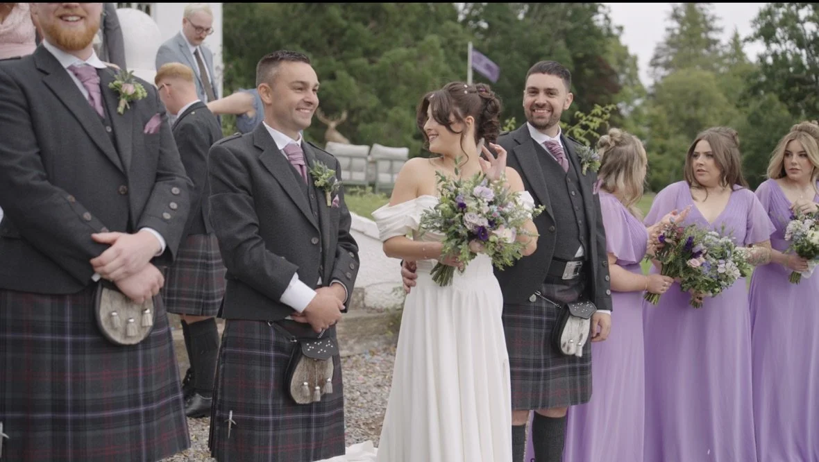 A group of people dressed in wedding attire at an outdoor wedding ceremony, with men in kilts and women in purple dresses, and a woman in a white wedding dress holding a bouquet of flowers.