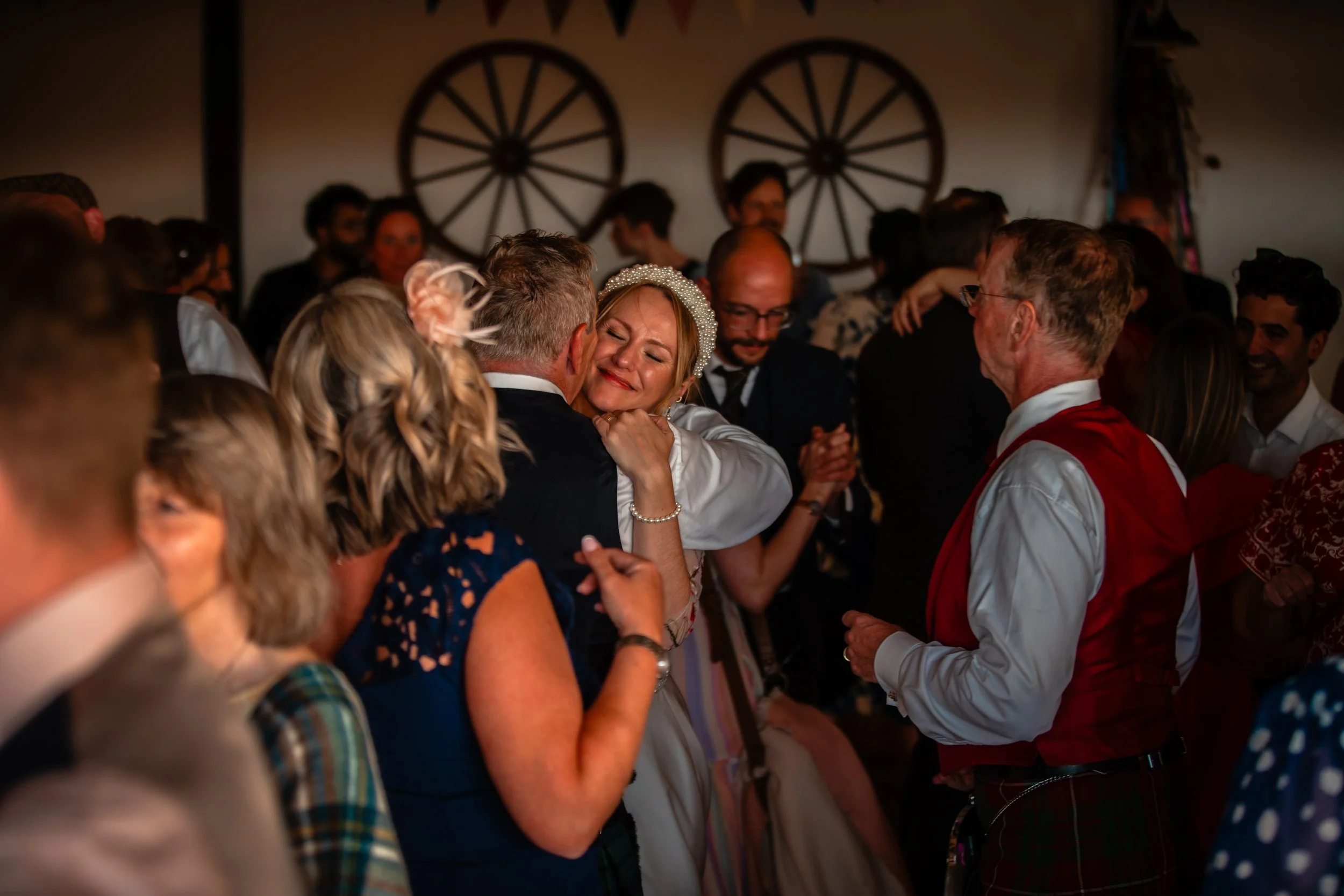 A woman in a white dress and pearl headband hugging a man amidst a crowd at a celebration, with others smiling and dancing in the background.