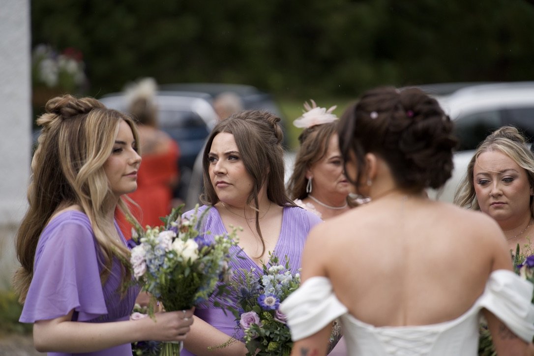 A group of women gathered outdoors, wearing purple and white dresses, holding bouquets, and appearing to converse, with cars and trees in the background. The woman in the foreground has her back to the camera.