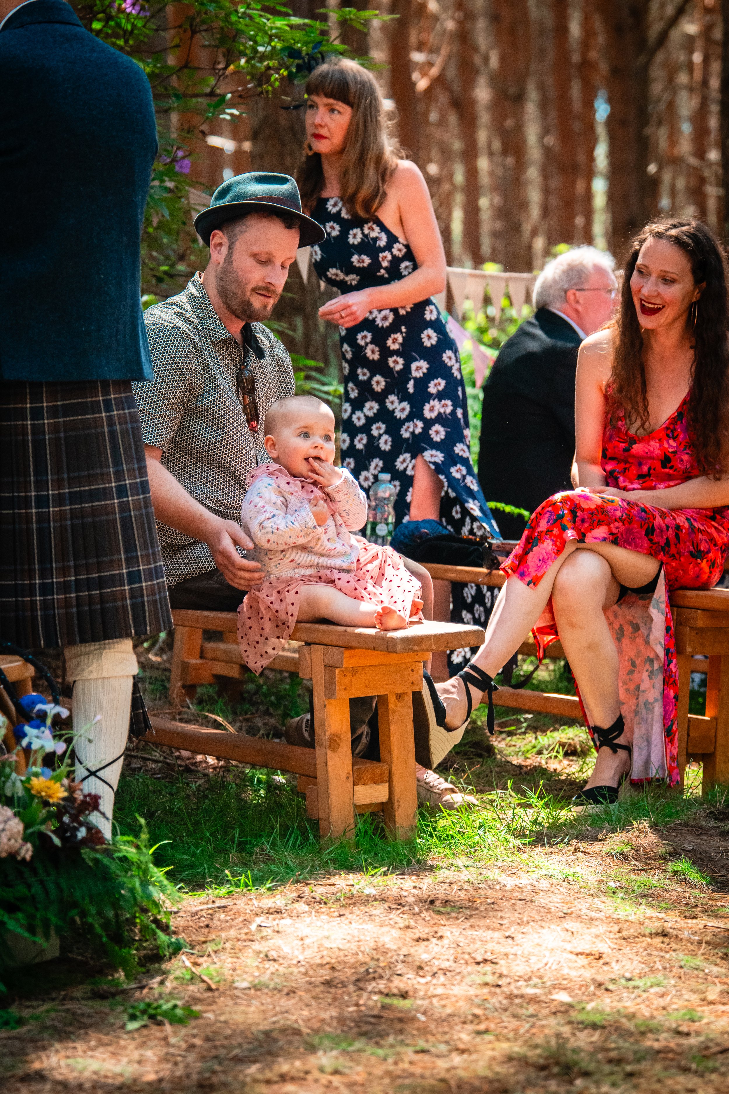 People at a forest gathering, a man with a hat sitting with a baby, a woman in a red floral dress laughing, and others chatting among trees.