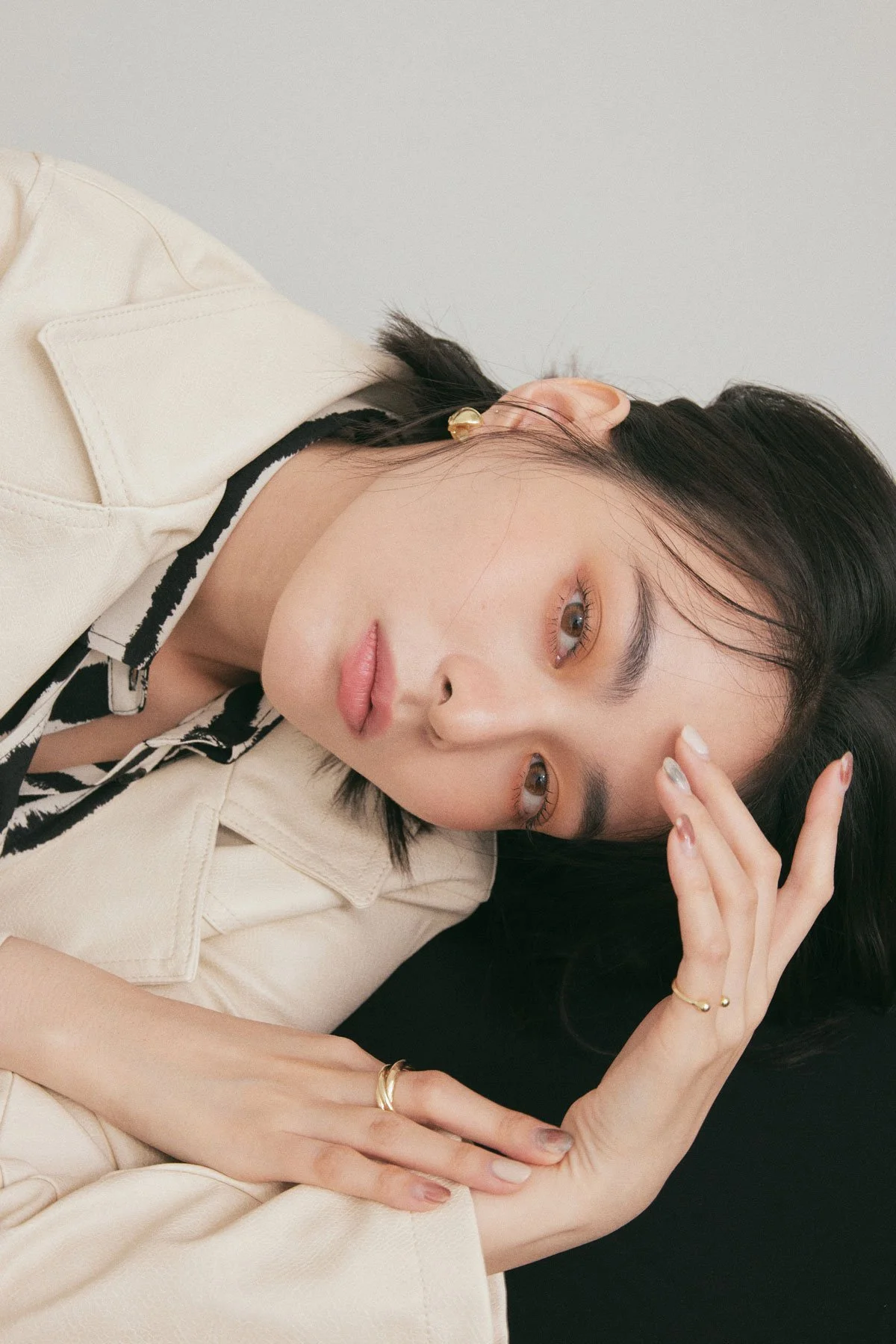 A woman with short dark hair lying on her side, wearing a beige jacket and black-and-white striped top, looking directly at the camera with a neutral expression.
