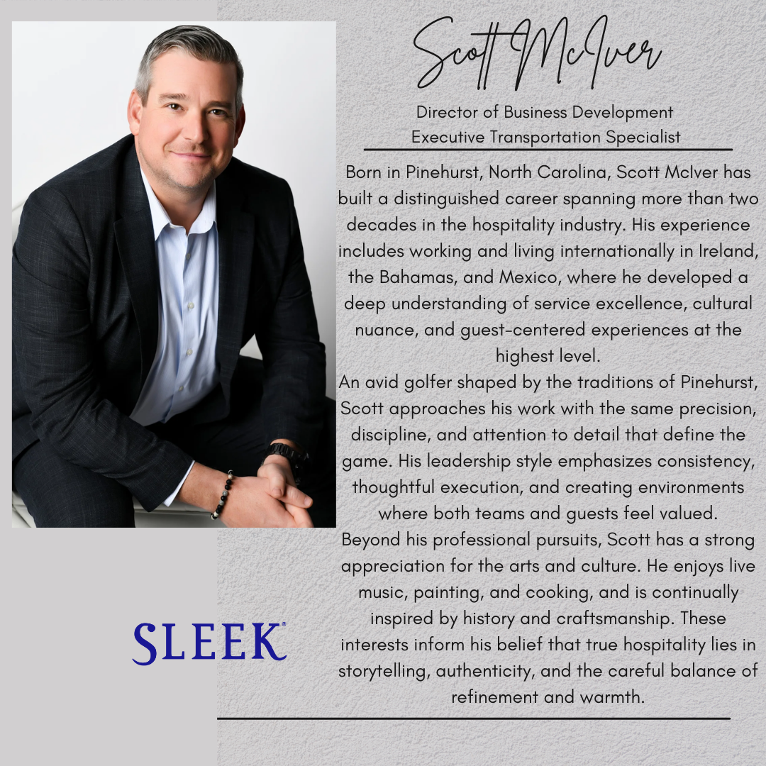 A man dressed in a dark suit and a light blue collared shirt, sitting with his hands clasped, smiling in a professional studio portrait.