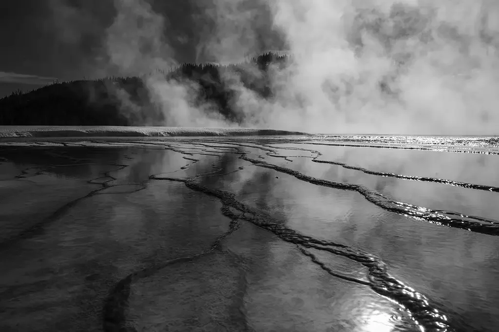 A black and white photo of a geothermal hot spring with steaming water, mineral deposits forming terraces, and a distant mountain range covered in clouds.