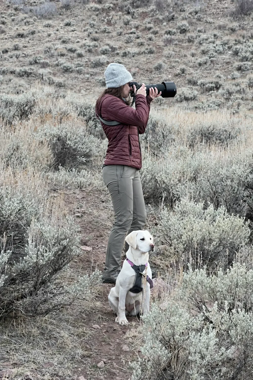 Rachel Albritton with a Nikon camera standing on a trail in the Flaming gorge area, a dry, shrub-covered landscape with a white Labrador Retriever sitting nearby.
