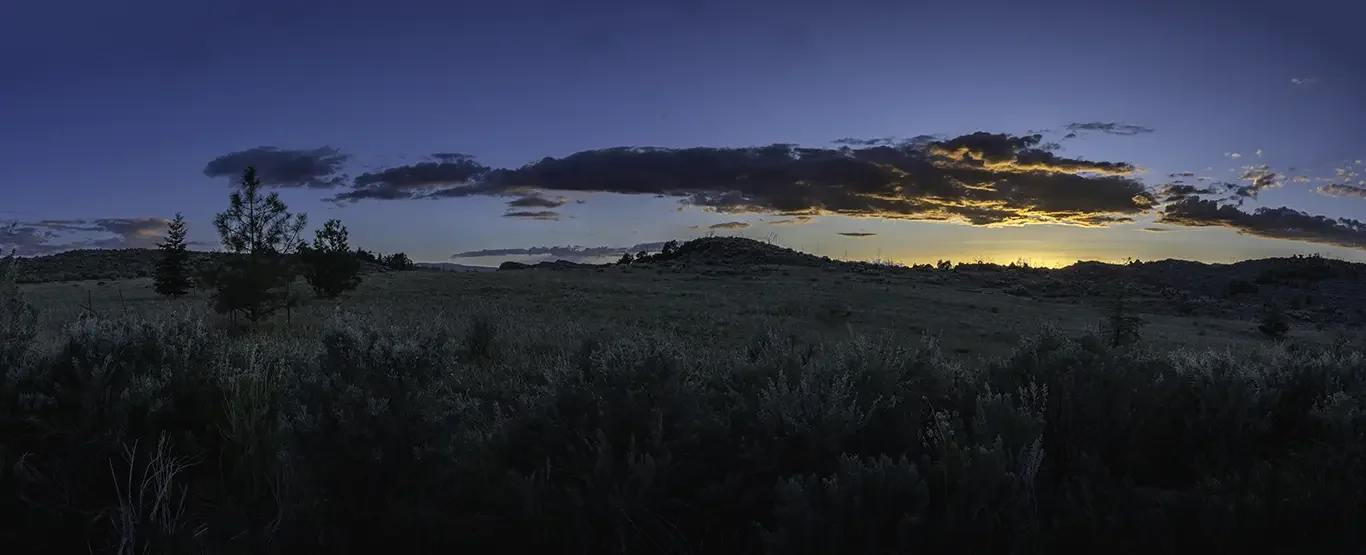 Sunset over a grassy field with scattered trees and hills in the distance, with clouds in the sky in Flaming Gorge / Dutch John, UT.