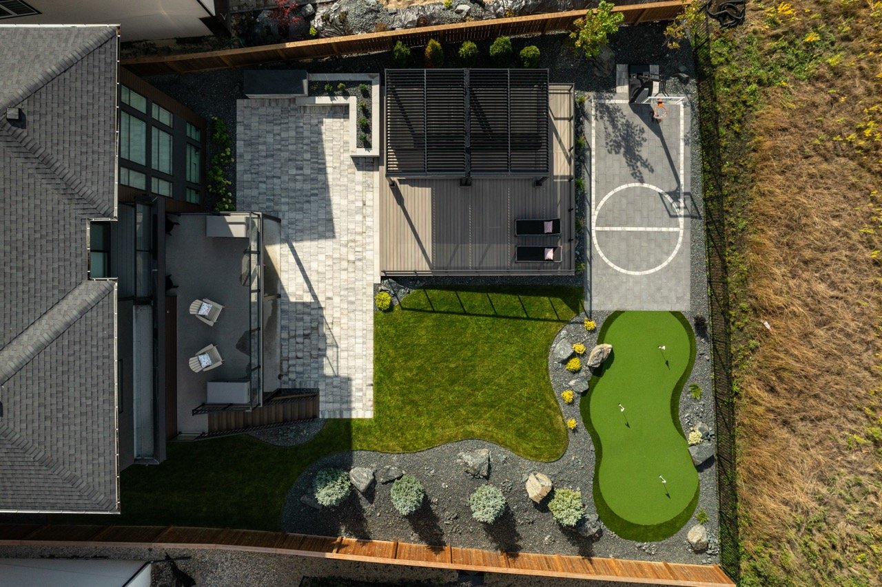 Aerial view of a backyard featuring a patio with seating, a lawn, a ping pong table, a small putting green, and a basketball court, all enclosed by a wooden fence.