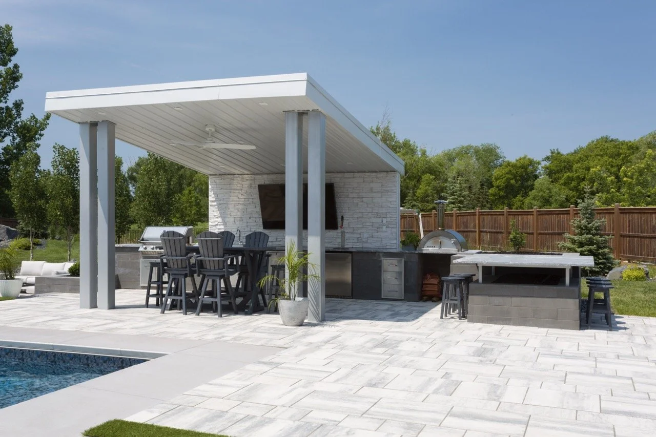 Outdoor kitchen with covered patio, dining table with high chairs, grill, pizza oven, and swimming pool.