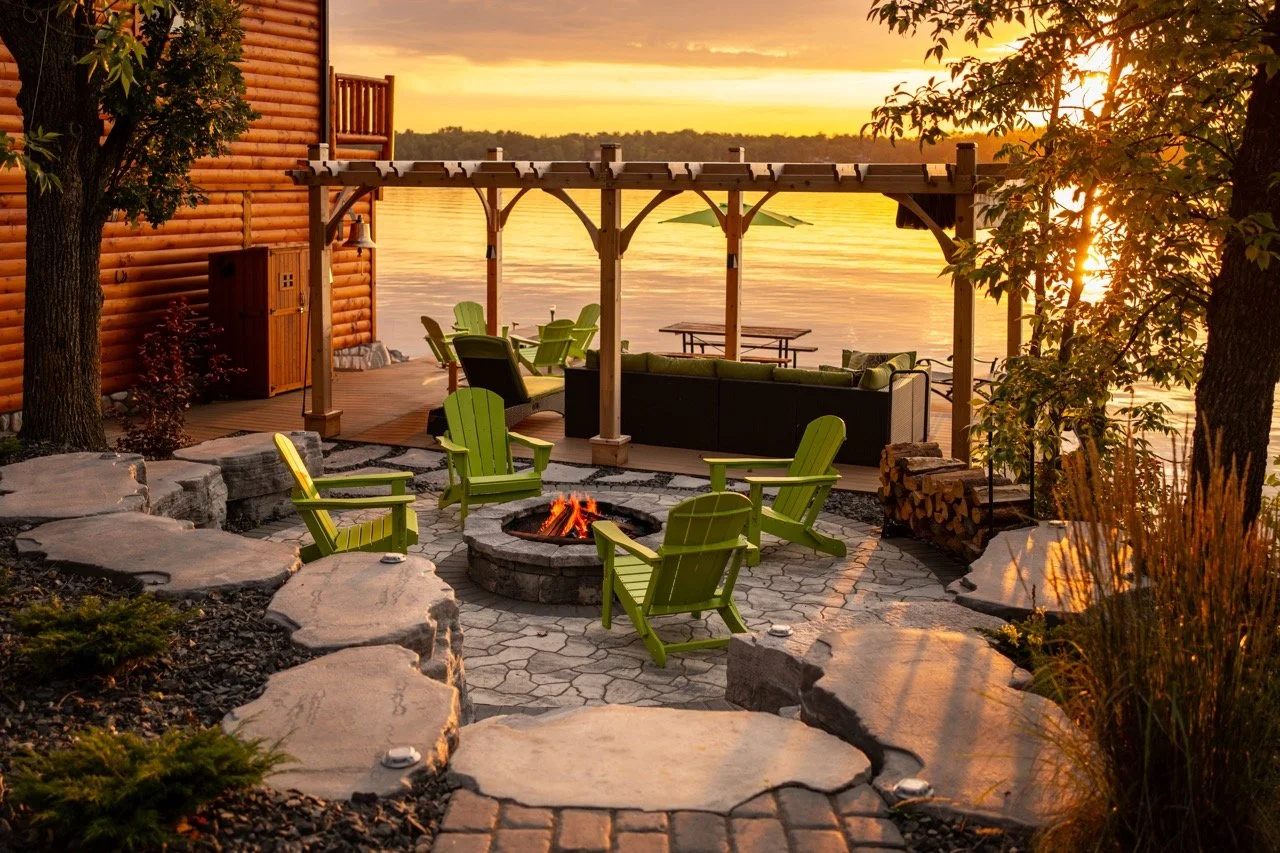 A cozy outdoor lakeside patio during sunset with a fire pit surrounded by green chairs, a wooden deck with lounge furniture under a pergola, and large rocks outlining a stone pathway, all beside a calm lake with a scenic sunset in the background.