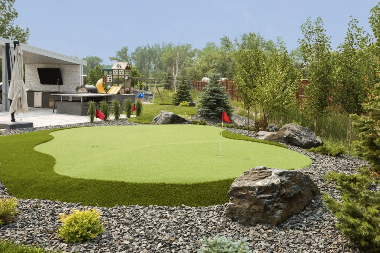 A backyard with a putting green, surrounded by rocks and shrubs. Nearby is a play area with a swing set and slide, next to an outdoor kitchen with a TV. Trees and a fence are in the background.