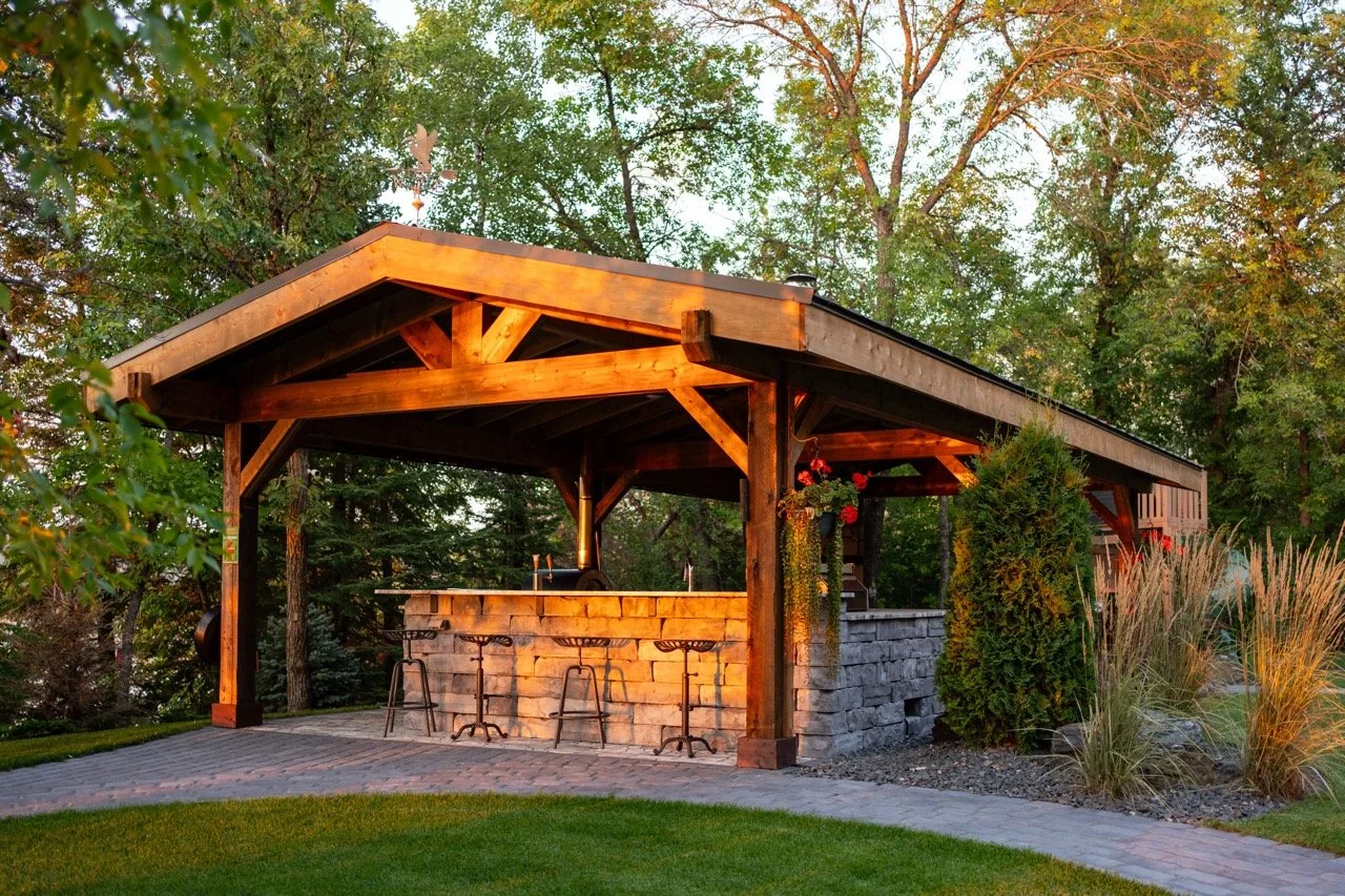 Outdoor covered patio area with bar counter and bar stools, surrounded by trees and landscaping at sunset.