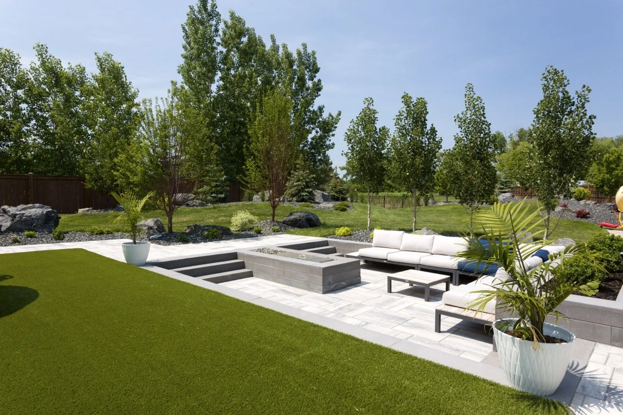 Modern backyard patio with a white sectional sofa, green lawn, potted plants, and surrounding trees.