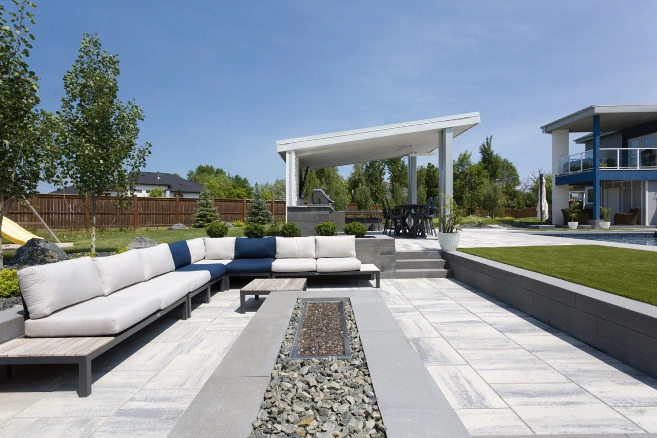 Modern backyard patio with a large white and blue sectional sofa, a fire pit with rocks, and a covered outdoor dining area. The space is surrounded by trees, a wooden fence, a slide, and well-maintained landscaping.