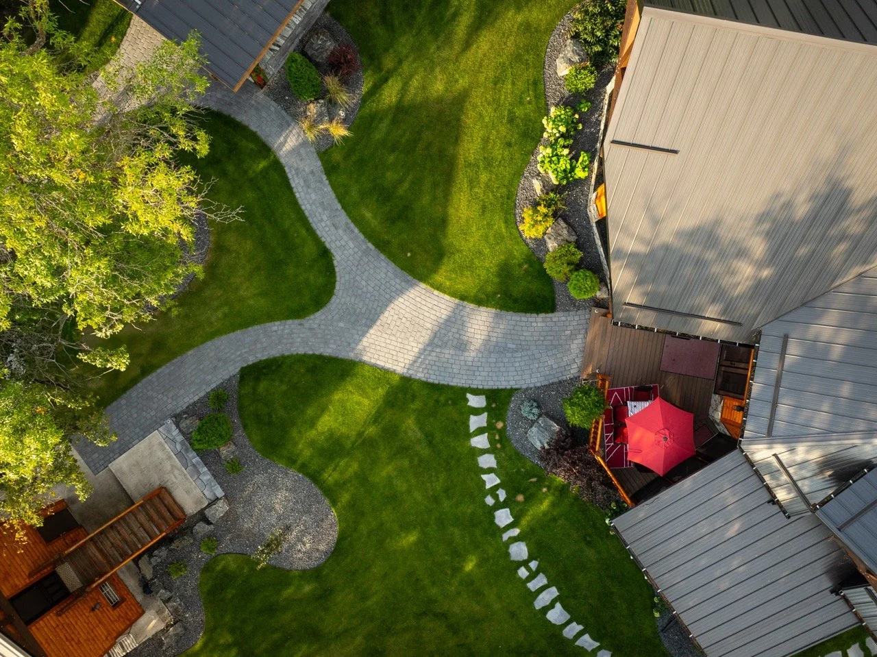 An aerial view of a backyard with a curved stone pathway, well-manicured green grass, trees, bushes, a red patio umbrella, and a wooden deck, surrounded by neighboring houses.