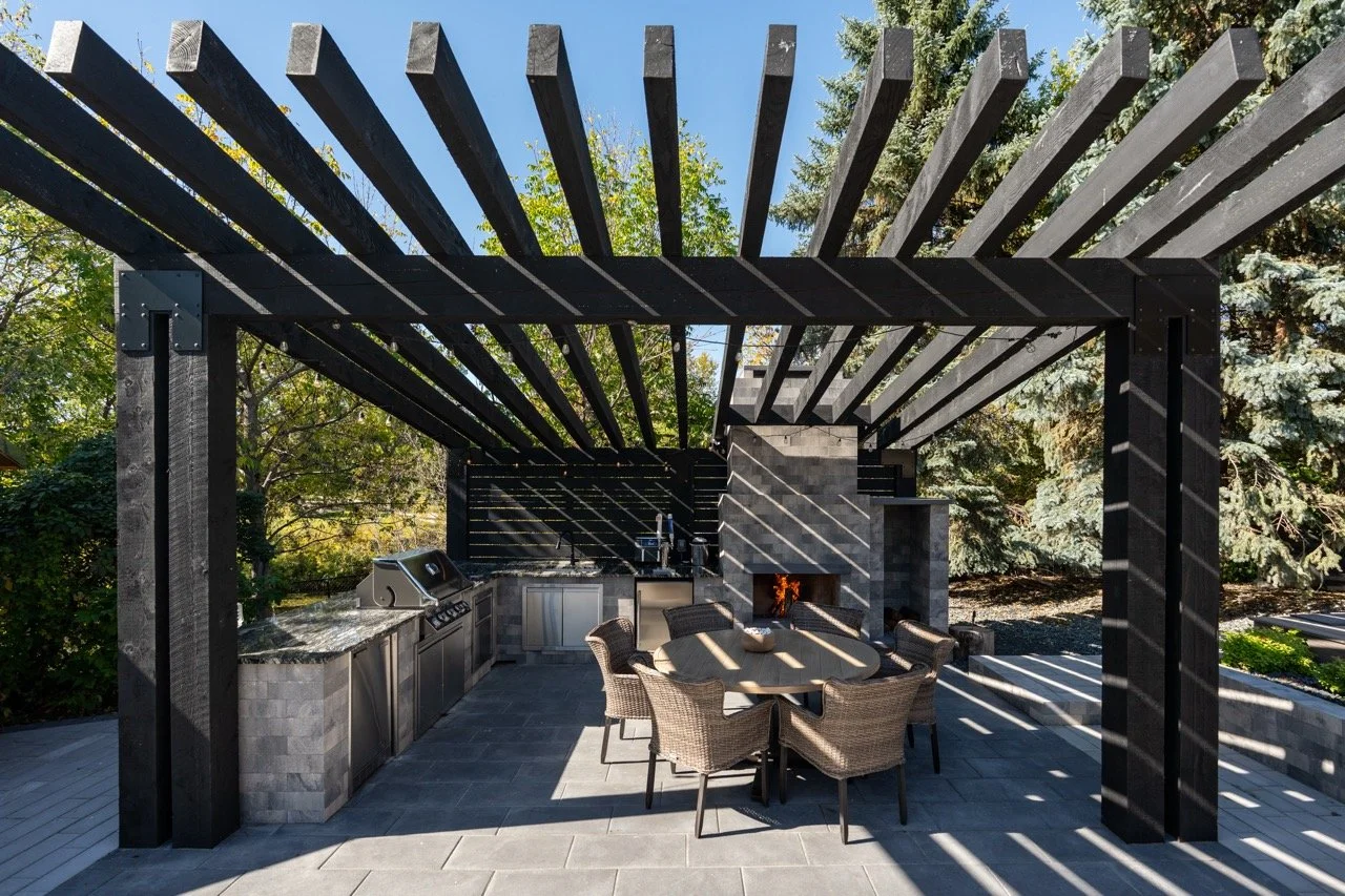 Outdoor kitchen and dining area with a black pergola, built-in grill, fire place, round table with six wicker chairs, surrounded by trees and blue sky.