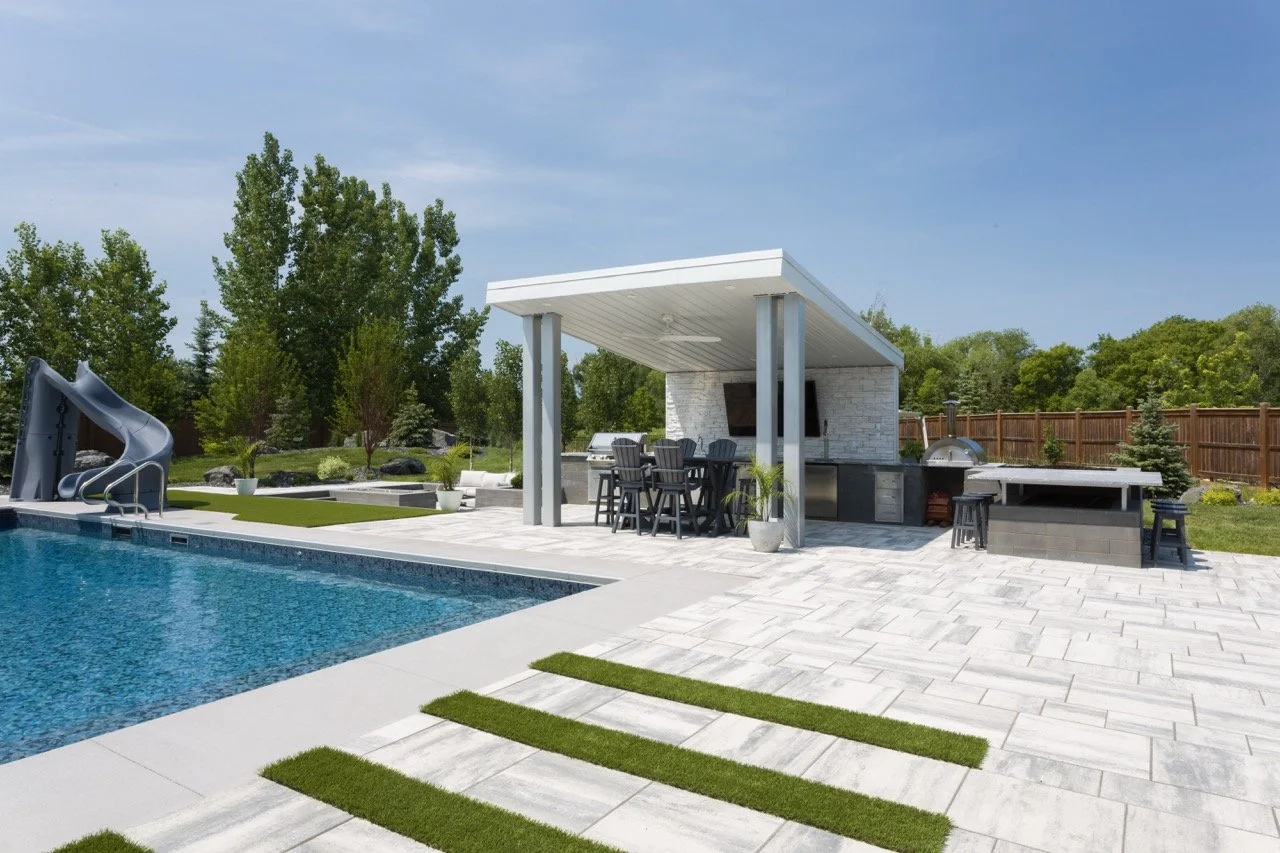 Backyard with pool, slide, patio, outdoor kitchen, and dining area under pergola, surrounded by trees and fencing.