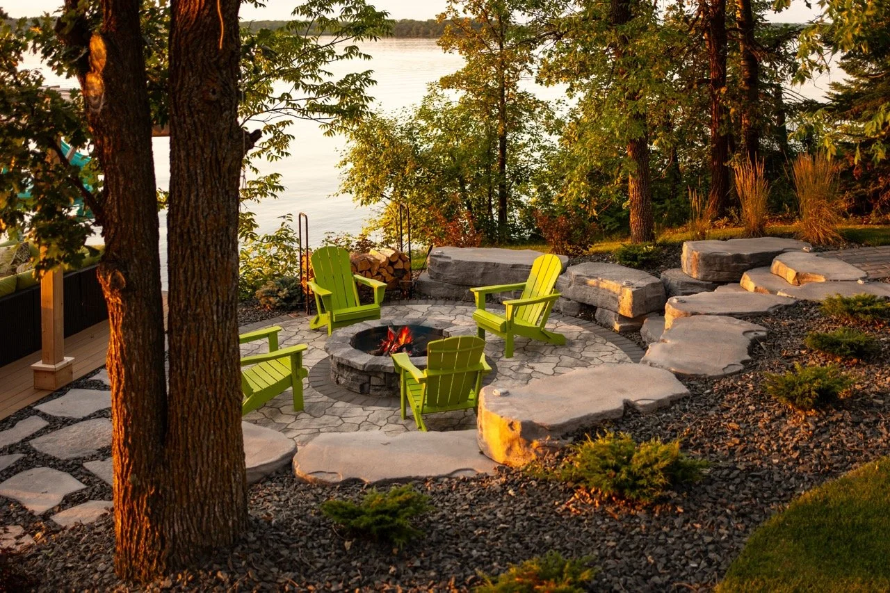 A backyard with a fire pit surrounded by four green Adirondack chairs on a stone patio, with large rocks, trees, and a view of a body of water in the background during sunset.