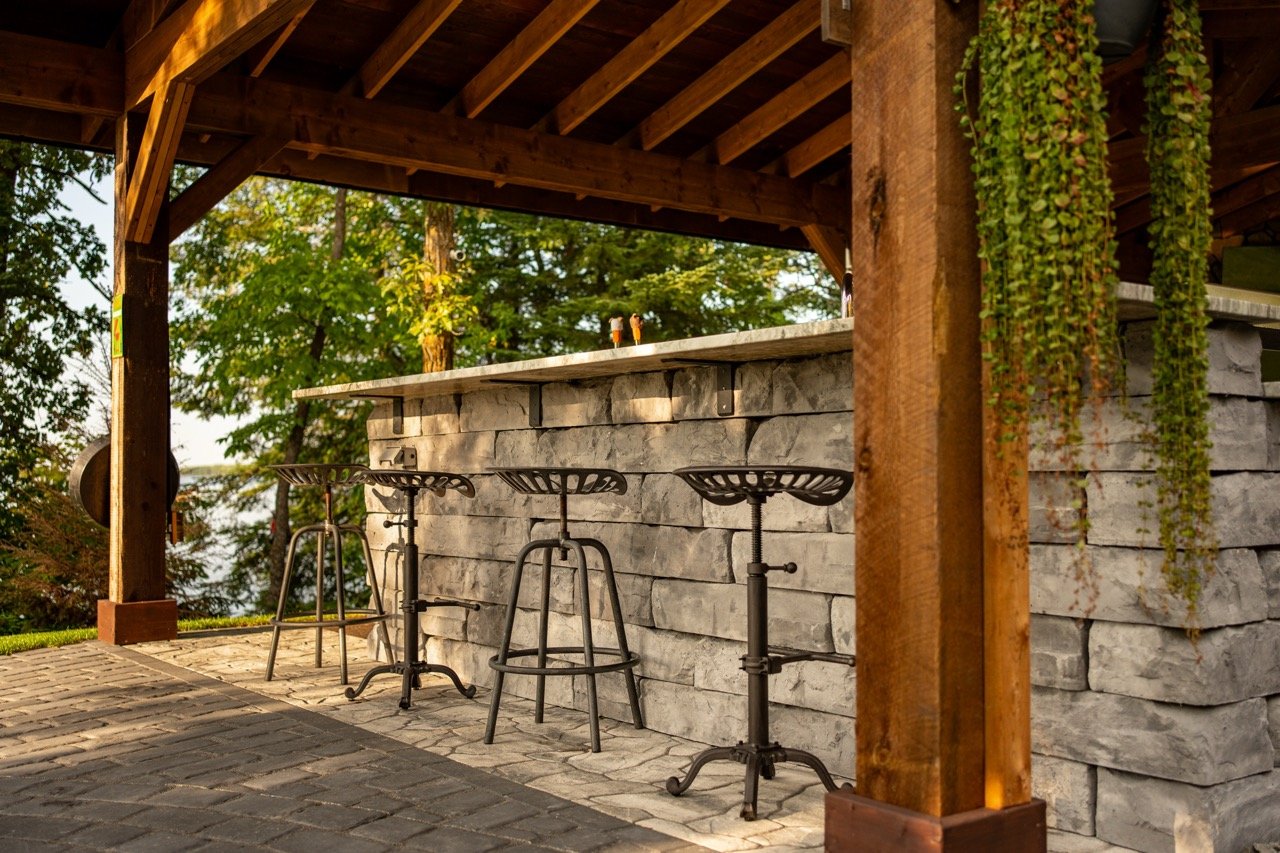 An outdoor bar area with a stone counter, three metal bar stools, and a wooden roof supported by wooden beams, surrounded by trees.