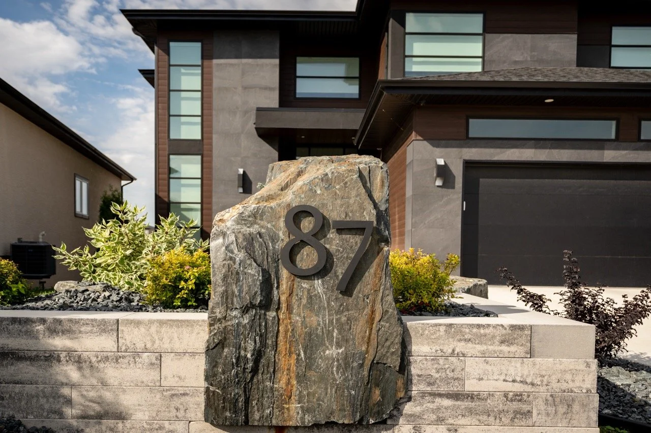 A modern house with a large stone plaque displaying the number 87, surrounded by landscaped plants and bushes, with a garage and windows visible on the house.