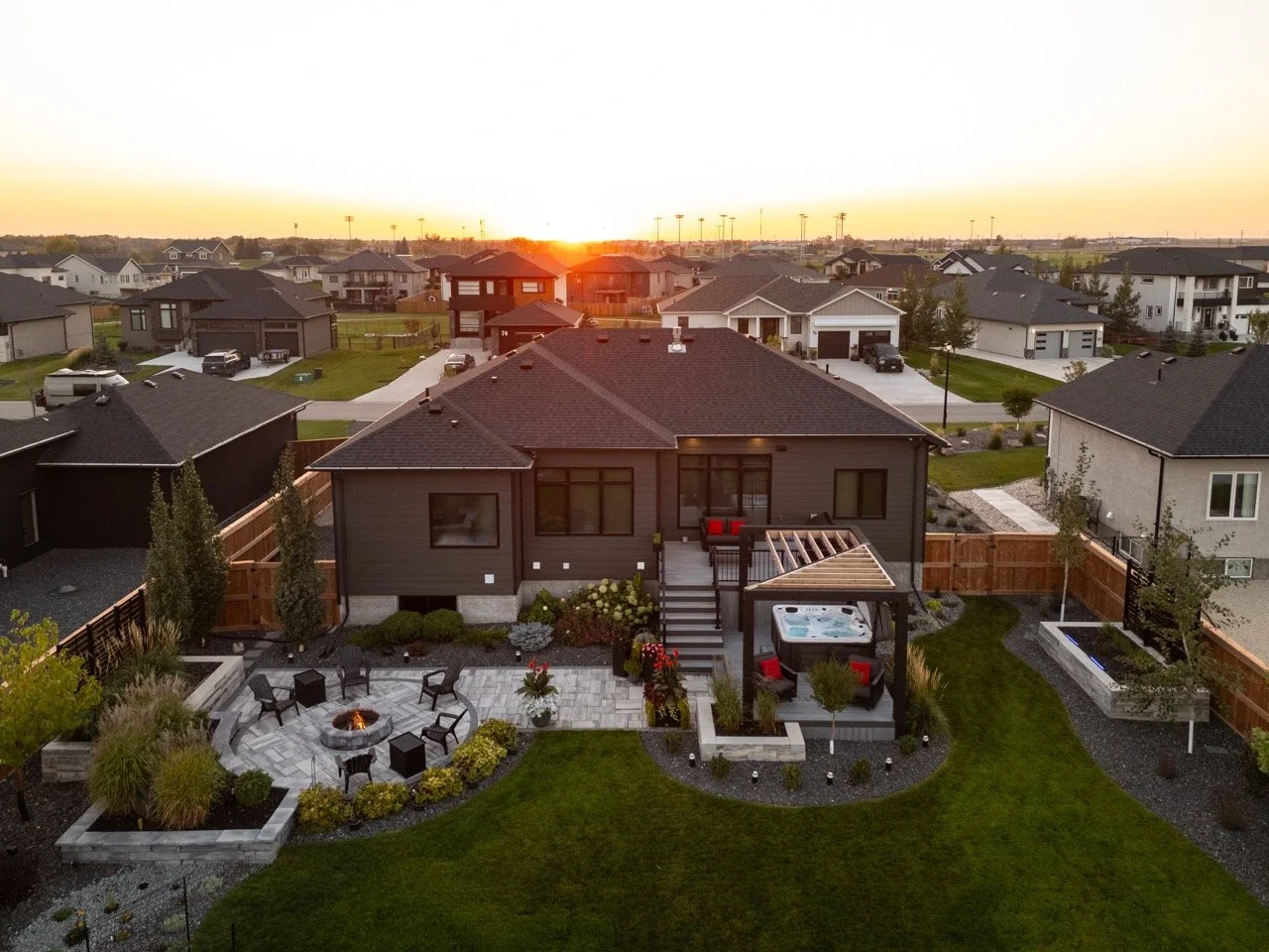Aerial view of a backyard with a fire pit, chairs, hot tub, and outdoor seating area during sunset, surrounded by other houses.