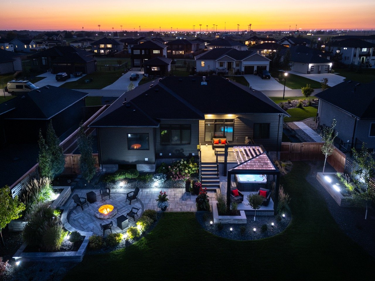 Aerial night view of a suburban neighborhood with illuminated houses, a backyard with a fire pit, outdoor seating, a hot tub, and landscaped garden beds.