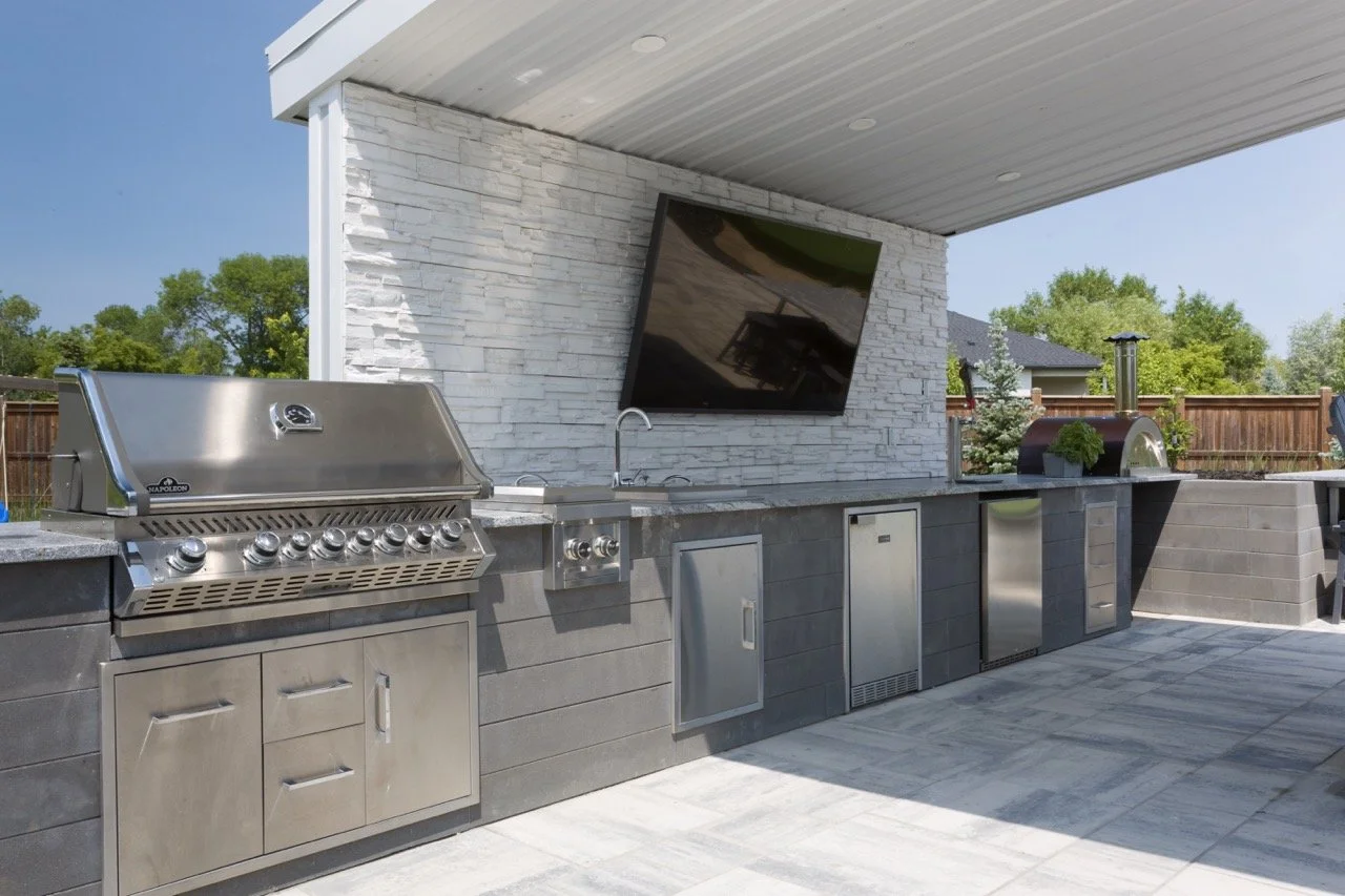 Outdoor kitchen with stainless steel grill, sink, and refrigerator under a pergola with a mounted TV.