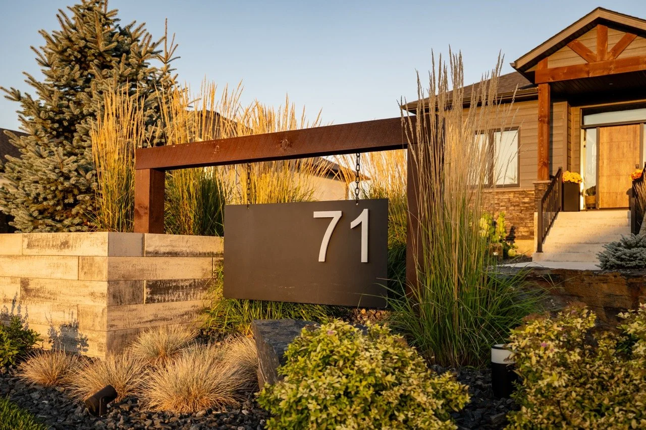 A modern house with the number 71 on a black sign in front of a landscaped yard with ornamental grasses and shrubs, a wooden fence, and a front porch with stairs.