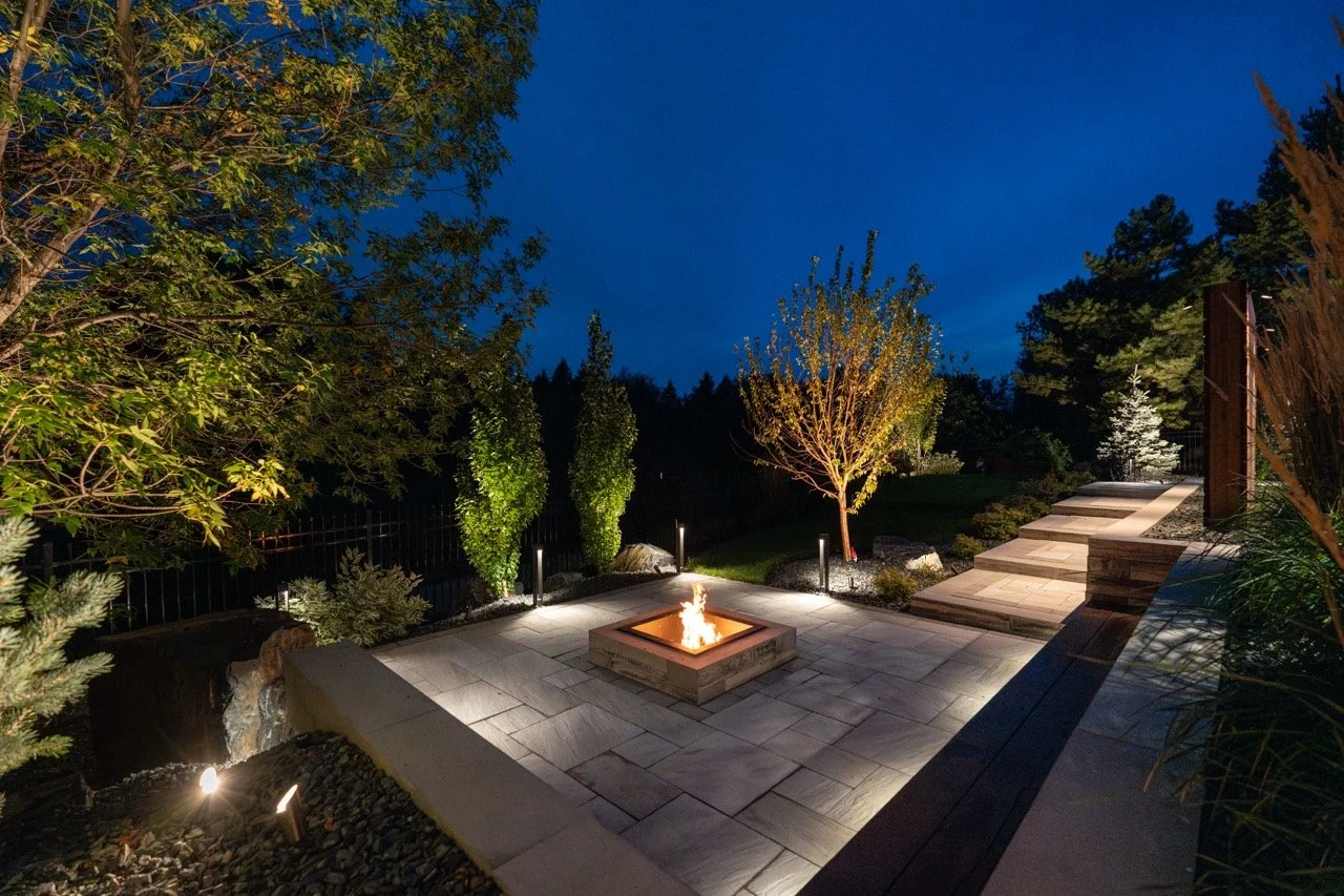 Outdoor patio at night with a lit fire pit, surrounded by trees and landscape lighting, adjacent to stone steps.