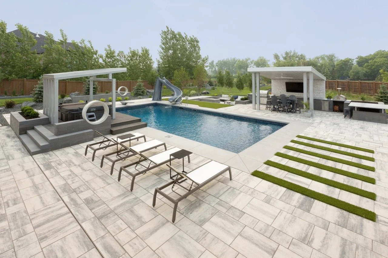 Modern backyard with swimming pool, lounge chairs, a slide, shaded cabana, and an outdoor dining area with kitchen. Surrounded by greenery and privacy fence.