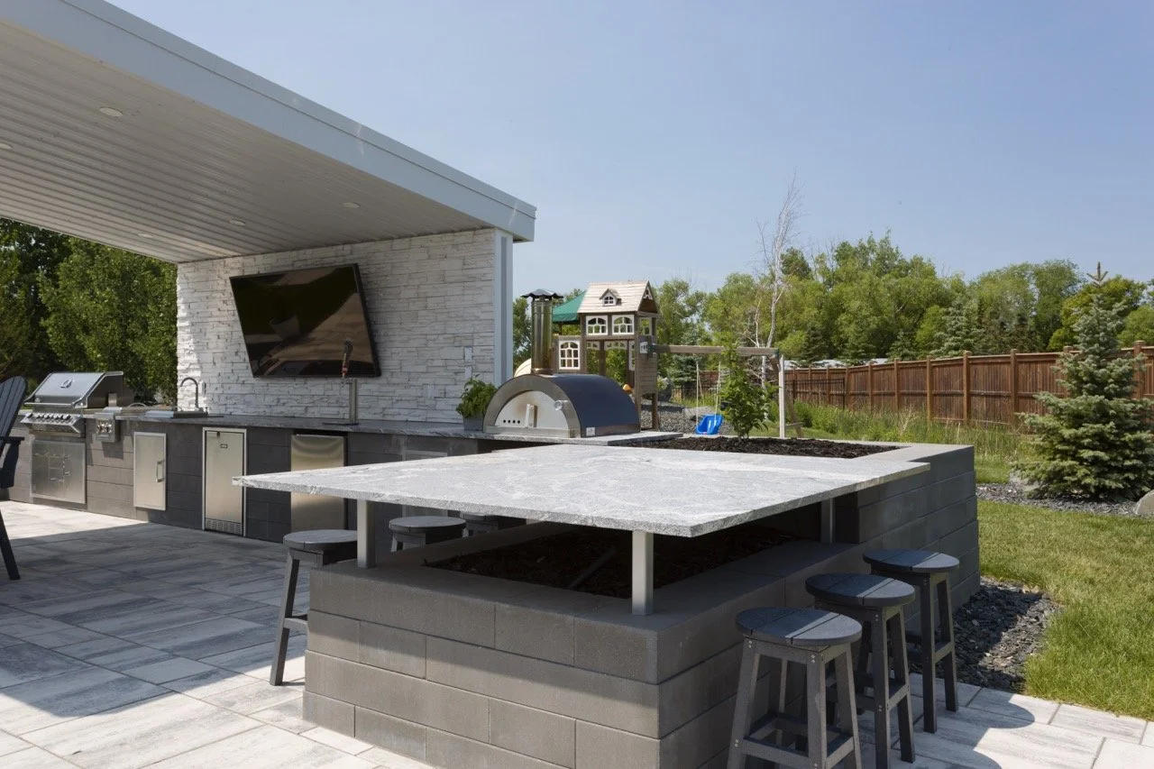 Modern outdoor kitchen with patio, featuring a grill, pizza oven, flat-screen TV, and a shaded countertop with stools, set in a landscaped backyard with a playhouse.