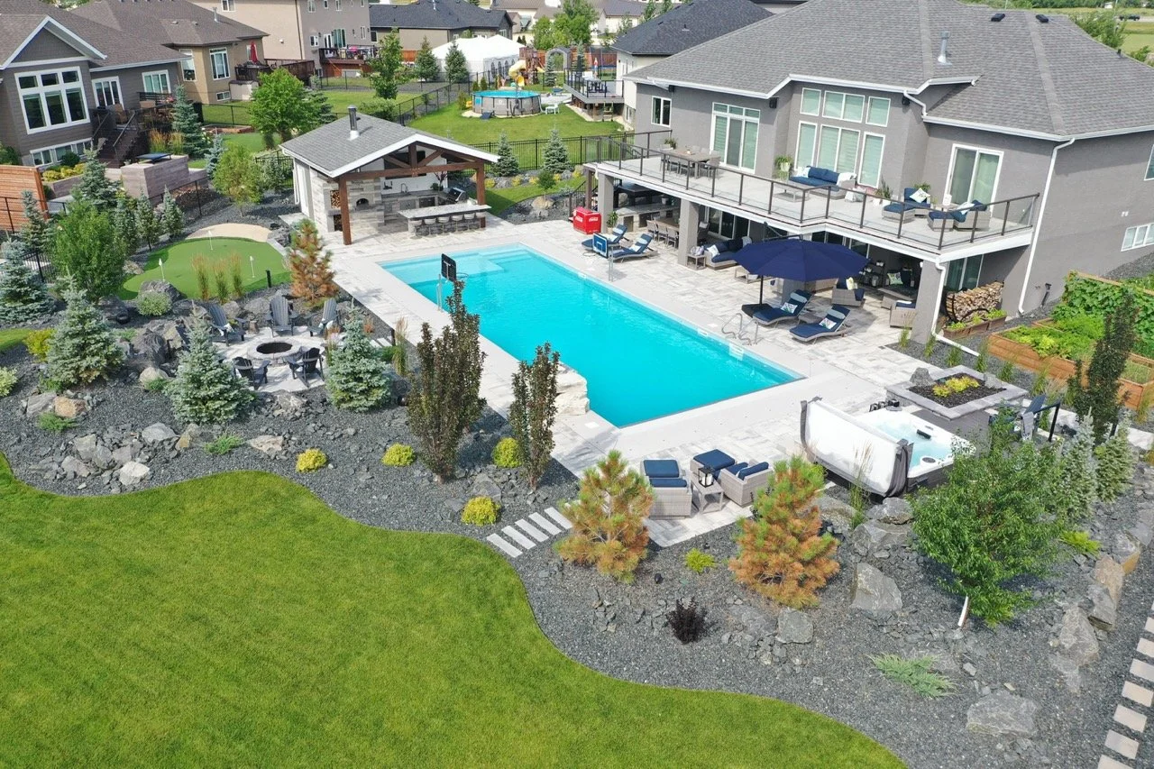 Aerial view of a backyard with a swimming pool, hot tub, outdoor seating, fire pit, and landscaped garden. Features a covered patio with an outdoor kitchen, surrounded by greenery and neighboring houses.