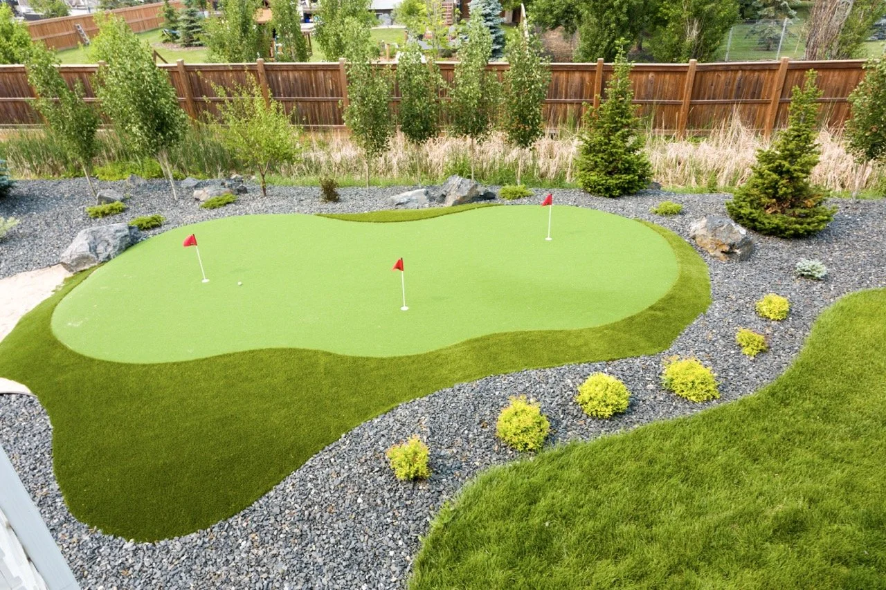 Backyard mini golf putting green with flags, surrounded by stones and landscaping, wooden fence in the background.