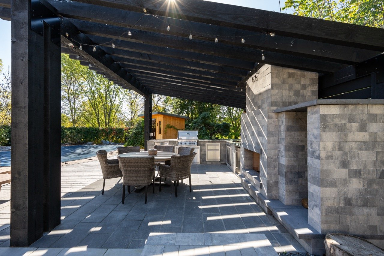 Outdoor patio with a large stone fireplace, a dining table with wicker chairs, a built-in grill, and string lights under a black pergola, surrounded by trees.