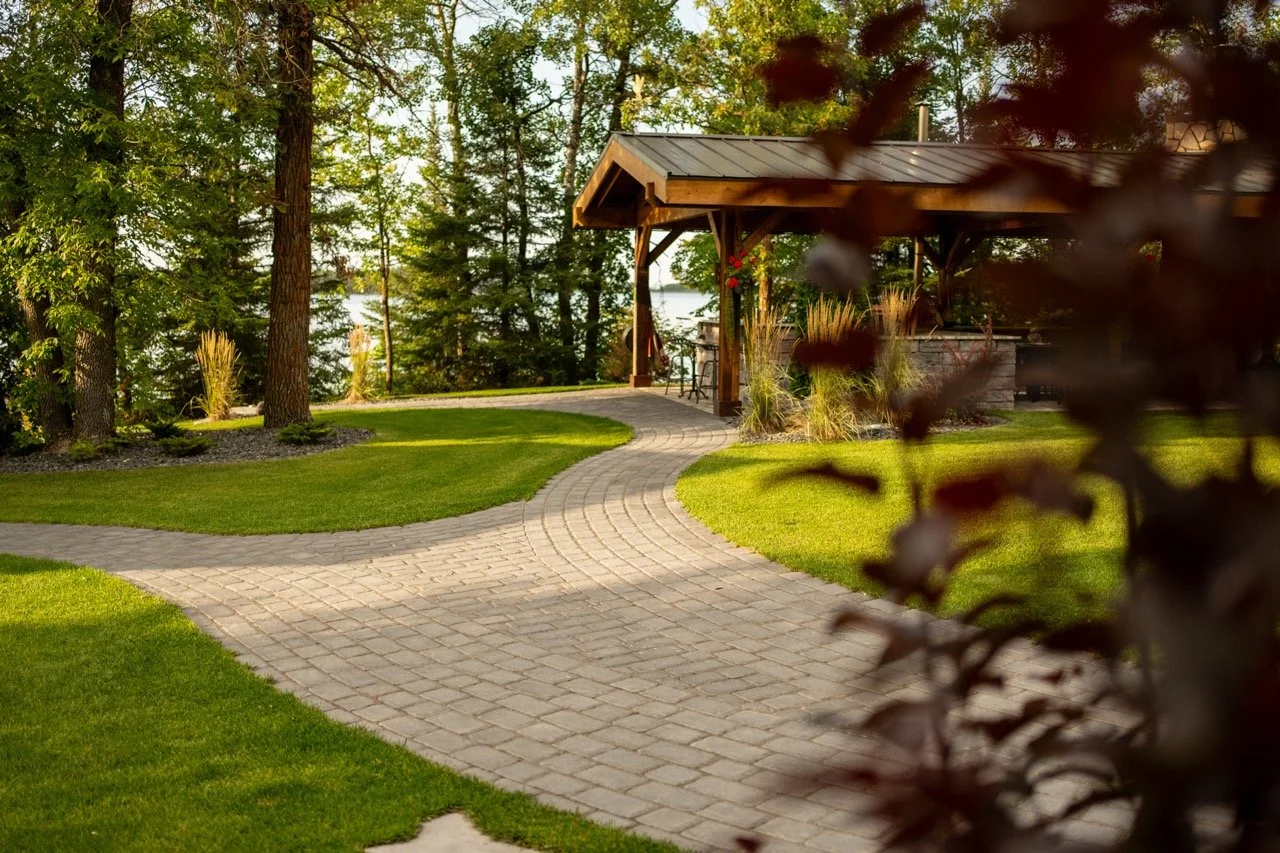 A paved walkway curves through a well-maintained lawn with trees and landscaping, leading to a wooden structure near a lake.