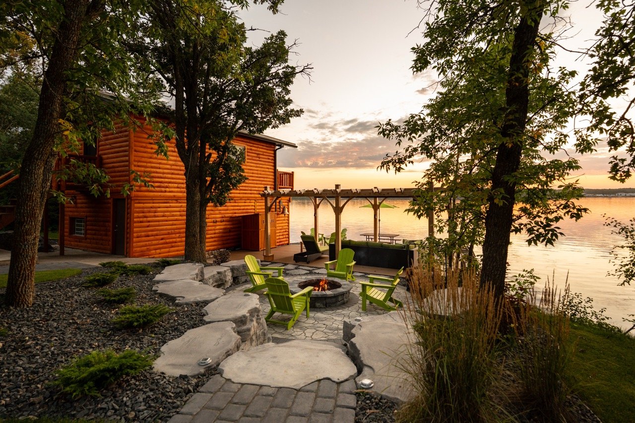 A lakeside backyard scene during sunset with a wooden cabin, green Adirondack chairs around a fire pit, on a stone patio, surrounded by trees and plants, overlooking the water.