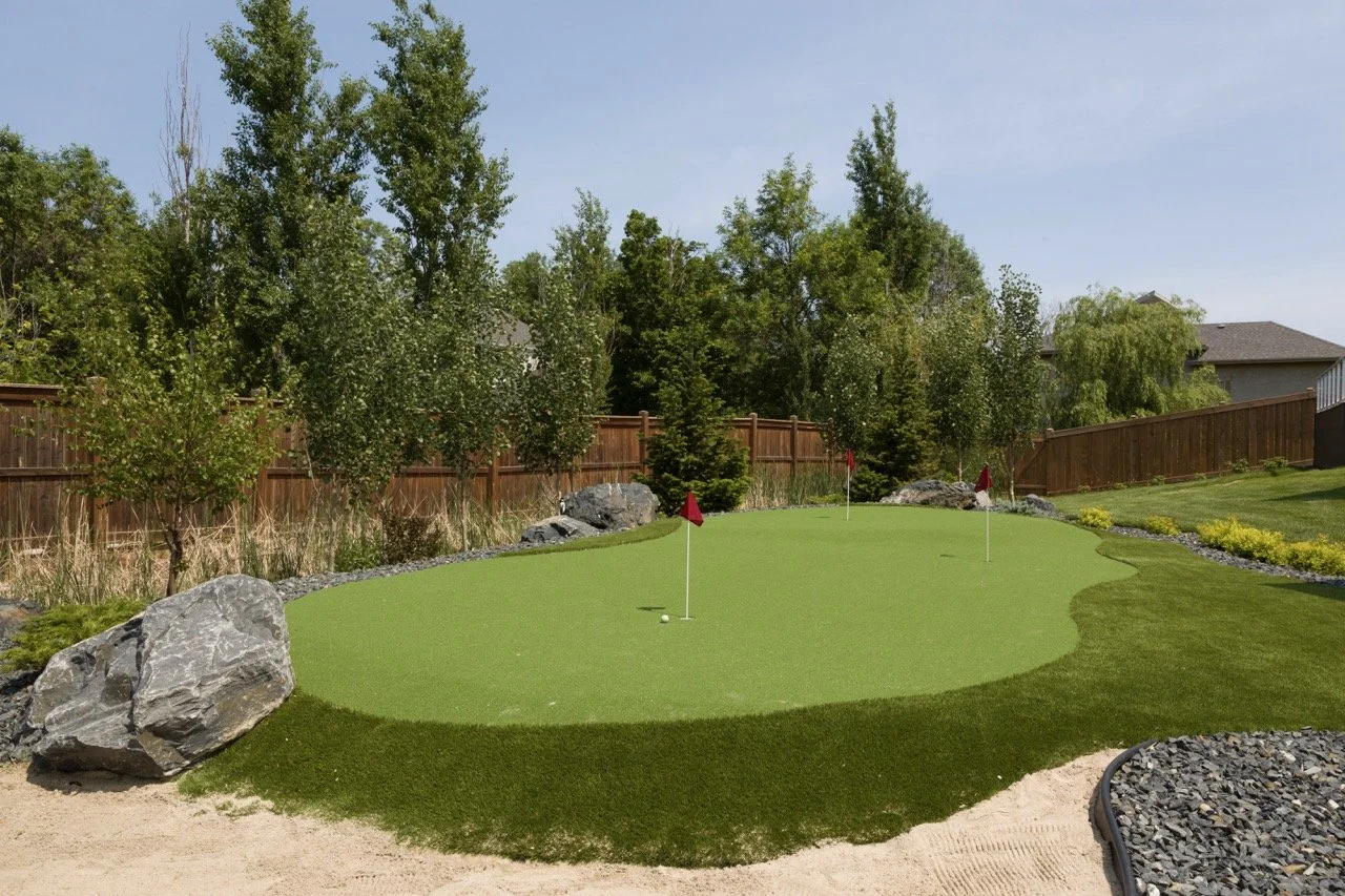 Backyard with a well-maintained artificial golf putting green, surrounded by greenery, rocks, and a wooden fence under a clear sky.