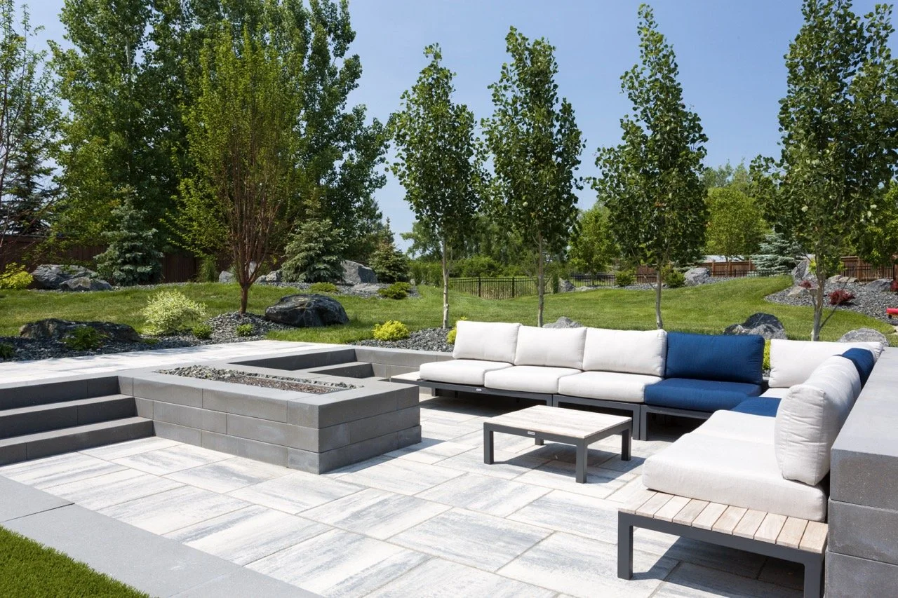 Outdoor patio with modern cushioned seating, stone fire pit, and surrounding lawn and trees.