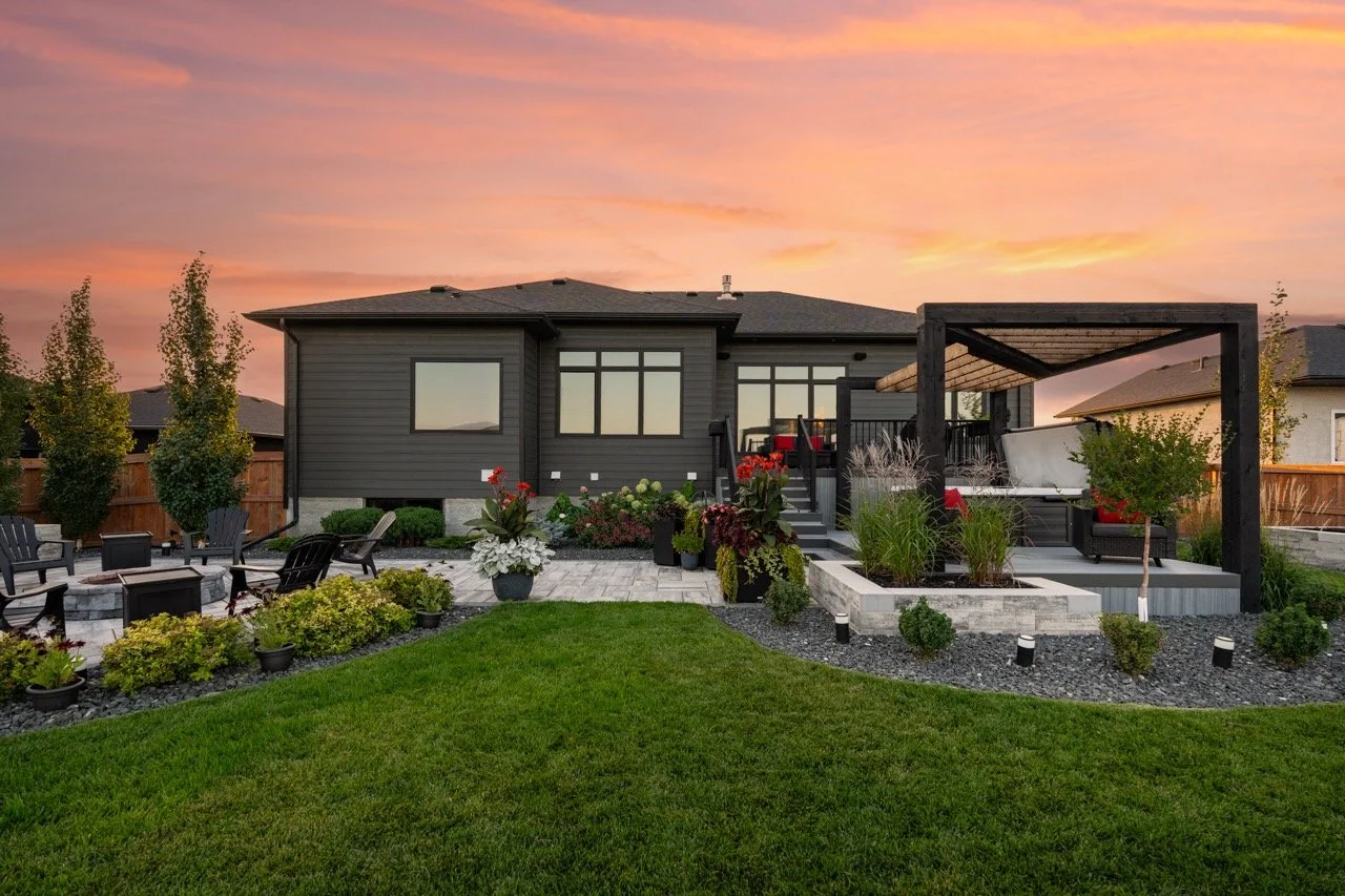 A backyard with a modern house in the background, featuring outdoor seating, a pergola with a swing, garden beds with plants and flowers, and a well-maintained lawn, all under a colorful sunset sky.